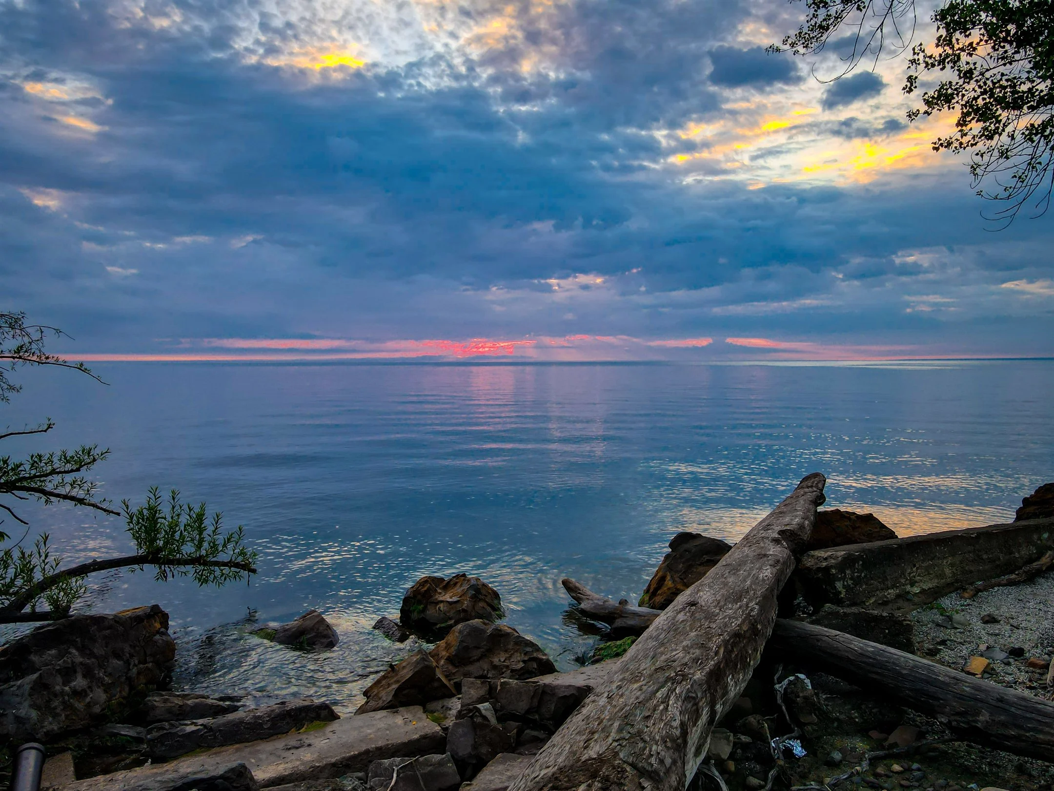 Scenic view of a serene lake with calm water and rocky shoreline at sunset. The sky is partly cloudy with hues of orange, pink, and blue. Foreground features a large driftwood log and some bushes.