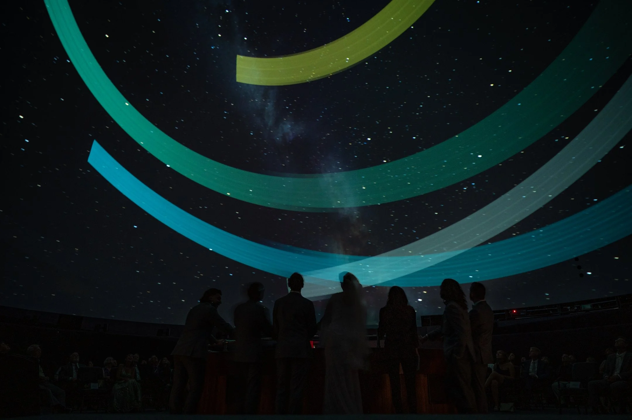 People inside a planetarium, stargazing with colored arcs projected on ceiling.