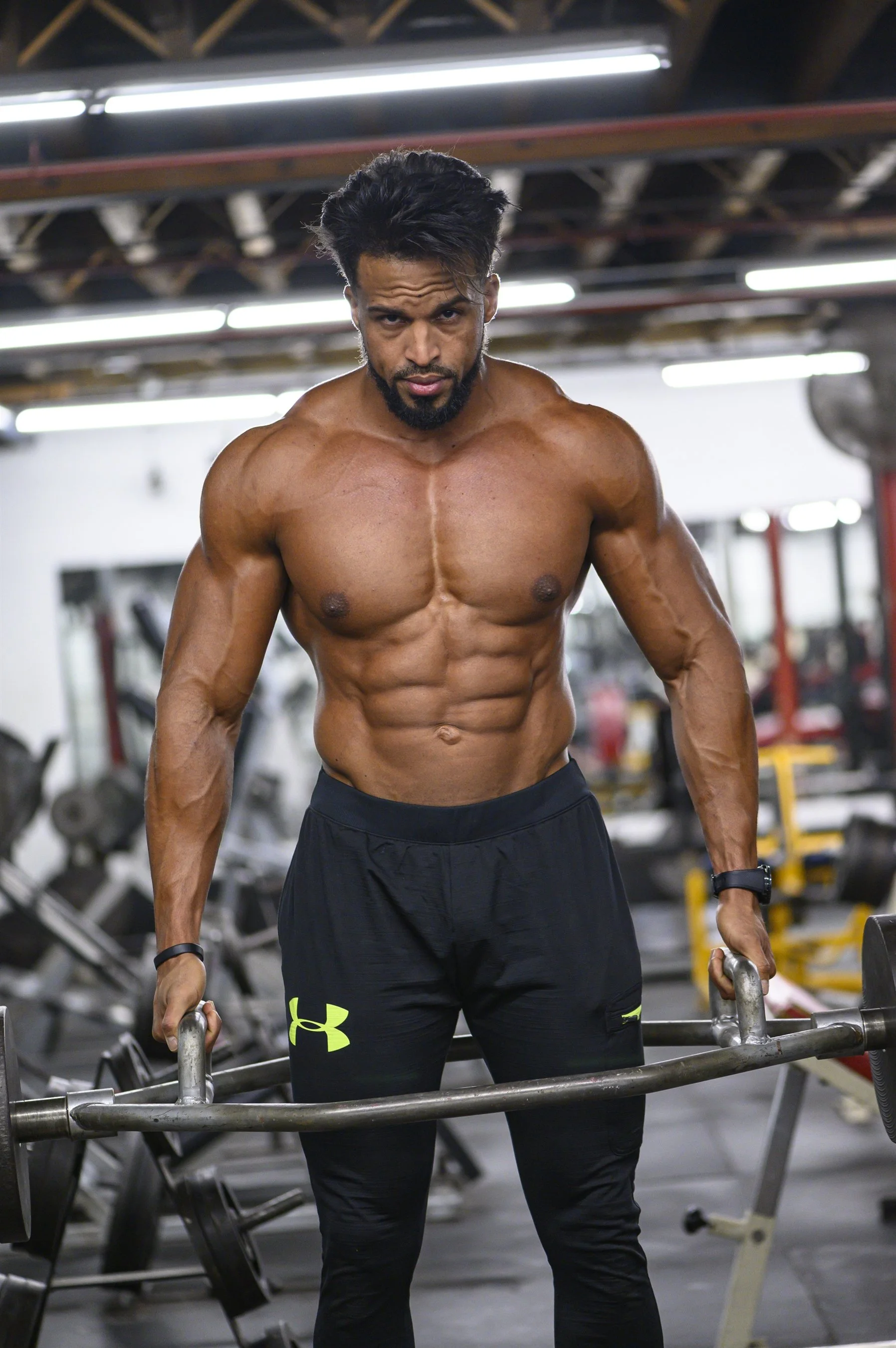 Muscular man lifting weights in a gym, wearing black pants, focused on exercise.