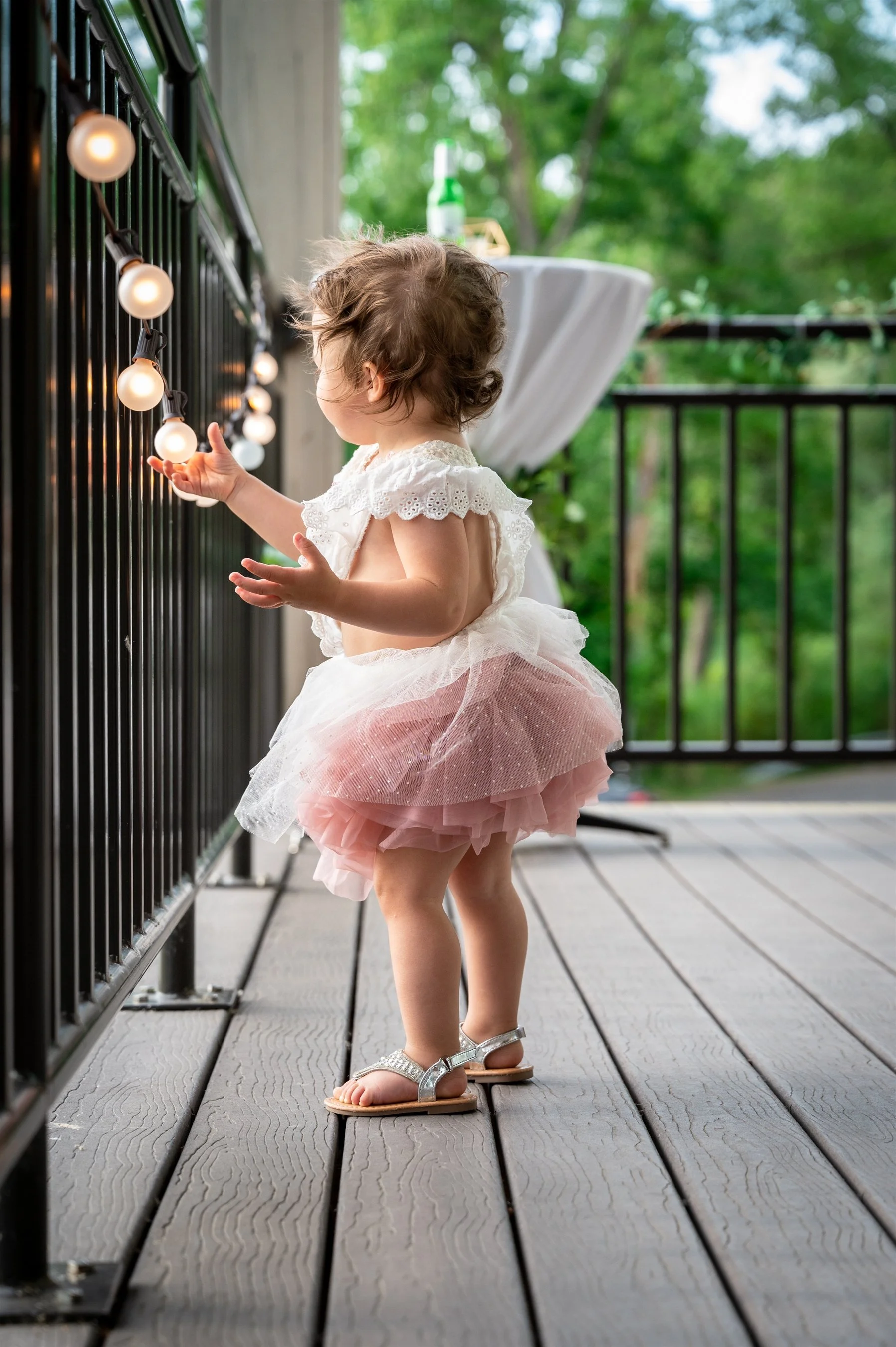 Toddler wearing a pink tutu and white shirt touches string lights on a porch.