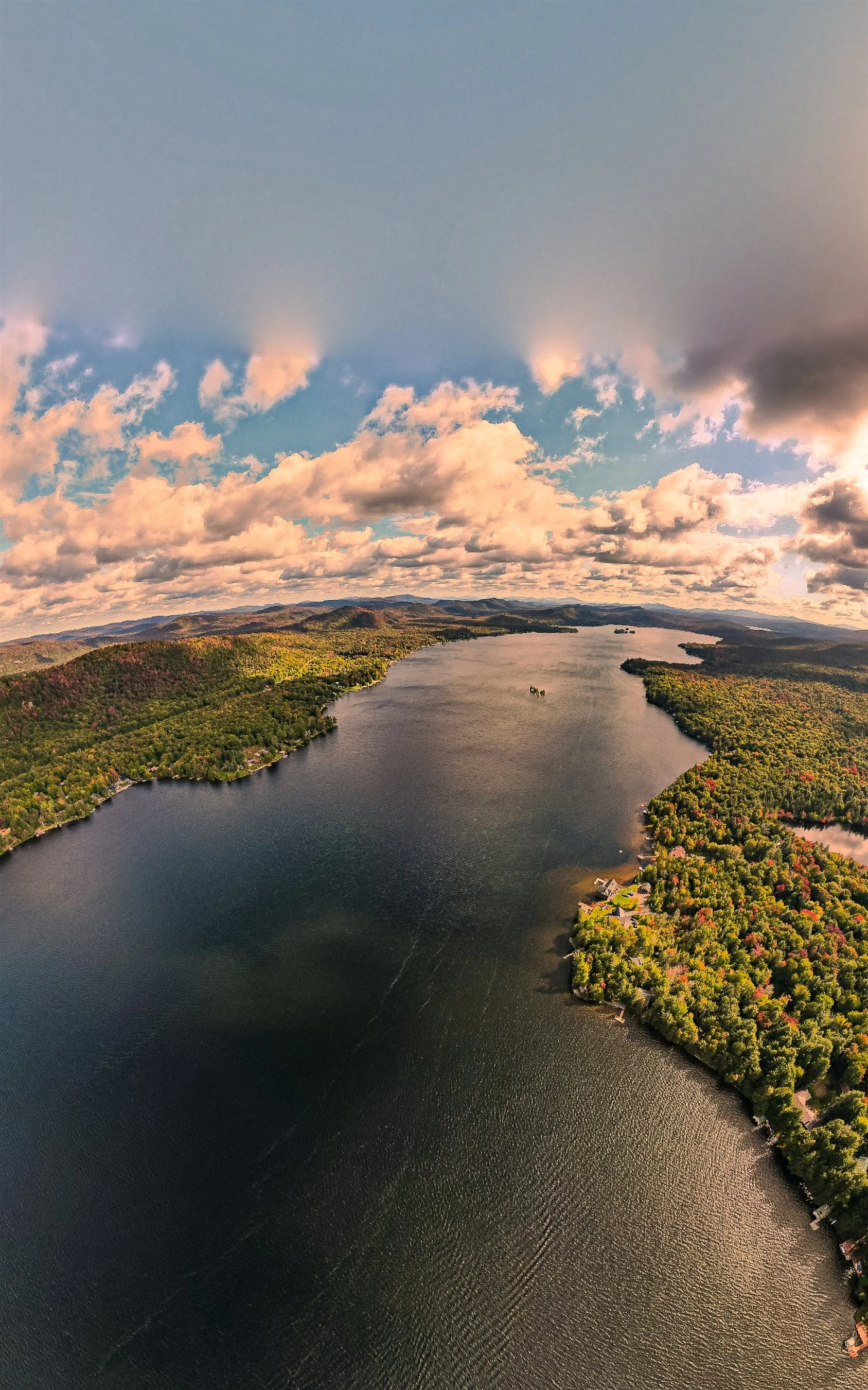 Aerial view of a large lake surrounded by lush green trees with autumn colors, under a partly cloudy sky.