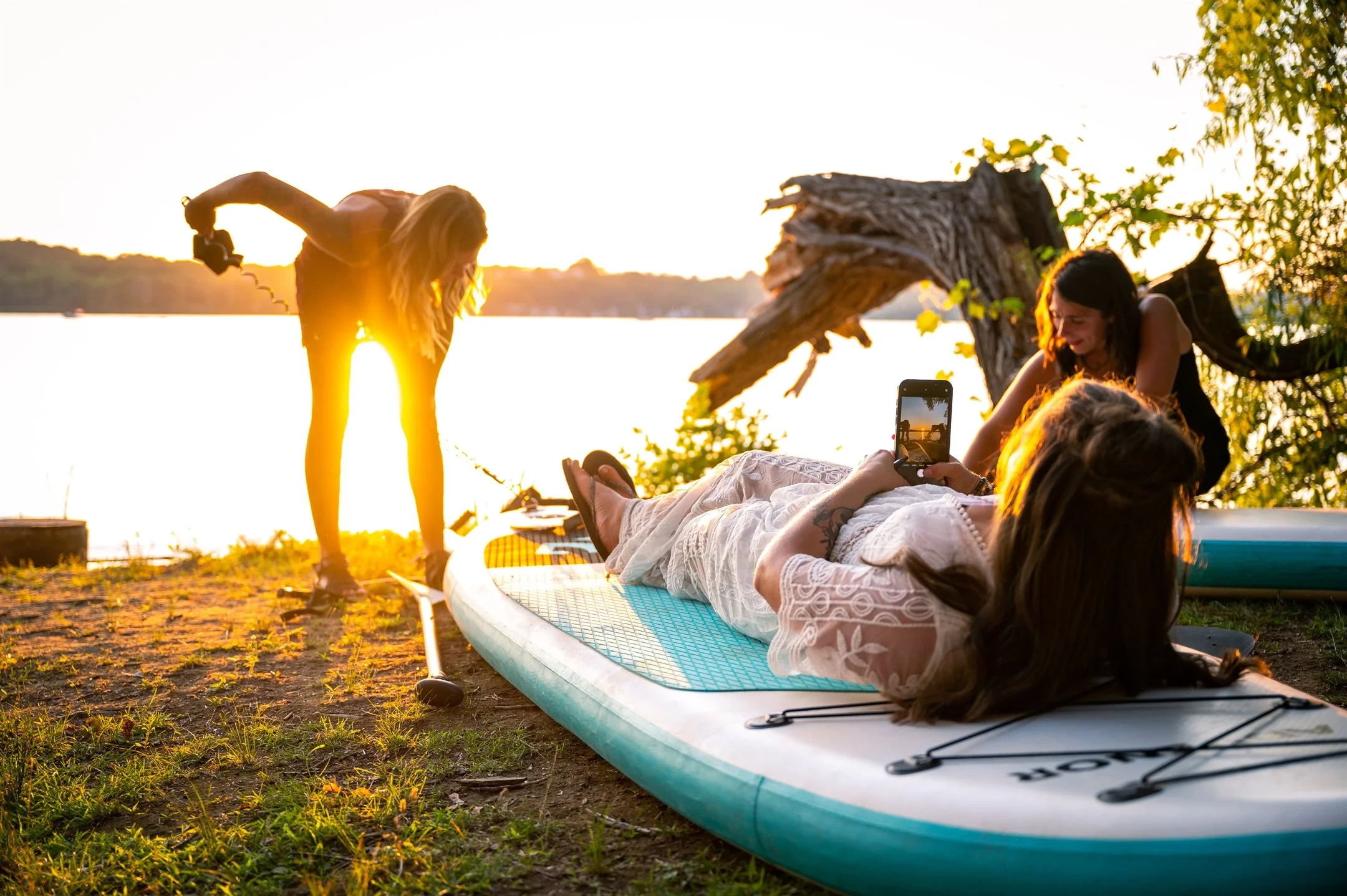 Sunset outdoor photoshoot with women near a lake, one laying on a paddleboard, capturing the moment with a smartphone.