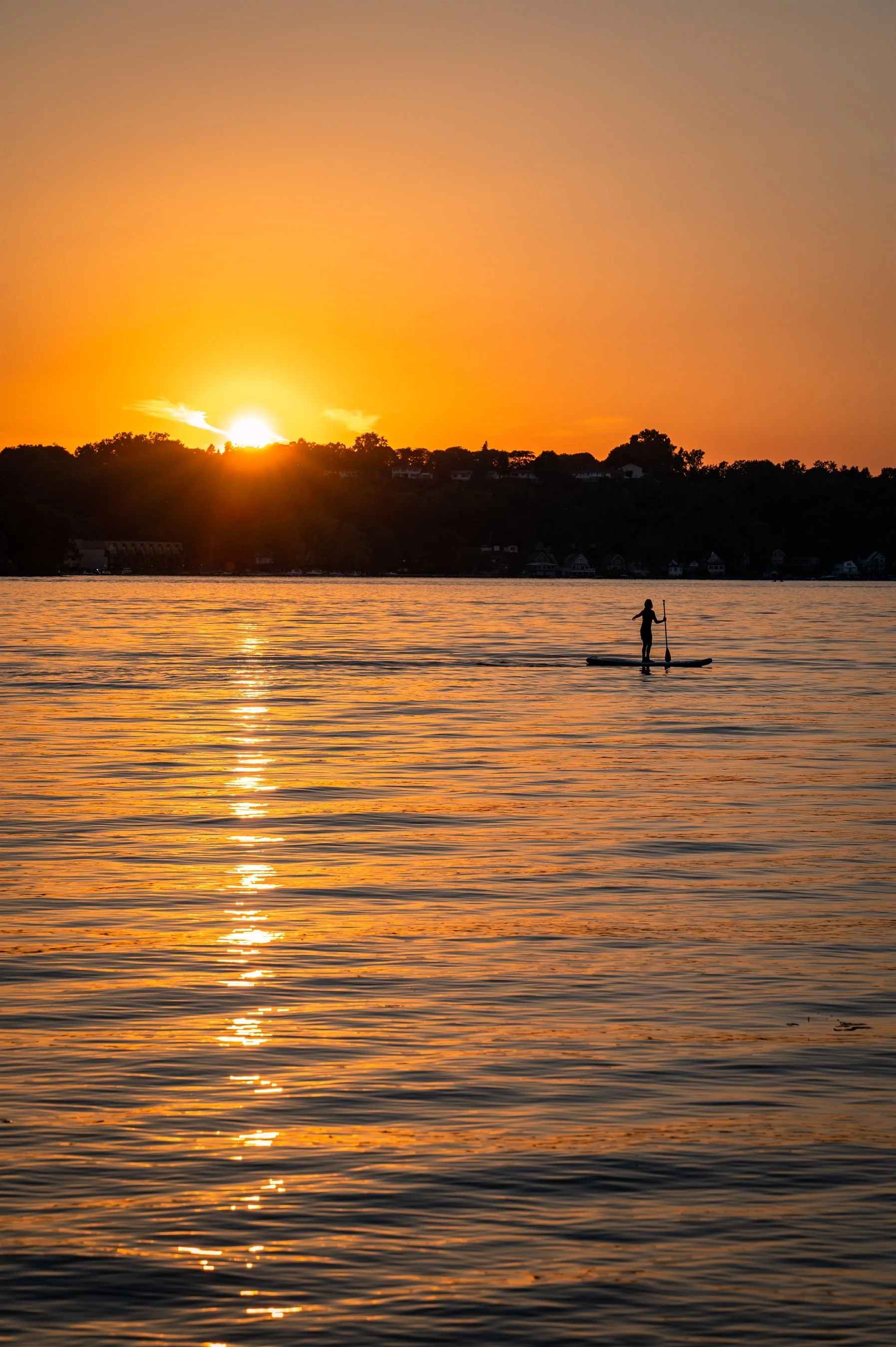 Silhouette of a person paddleboarding on a lake during sunset with an orange sky reflecting on the water.