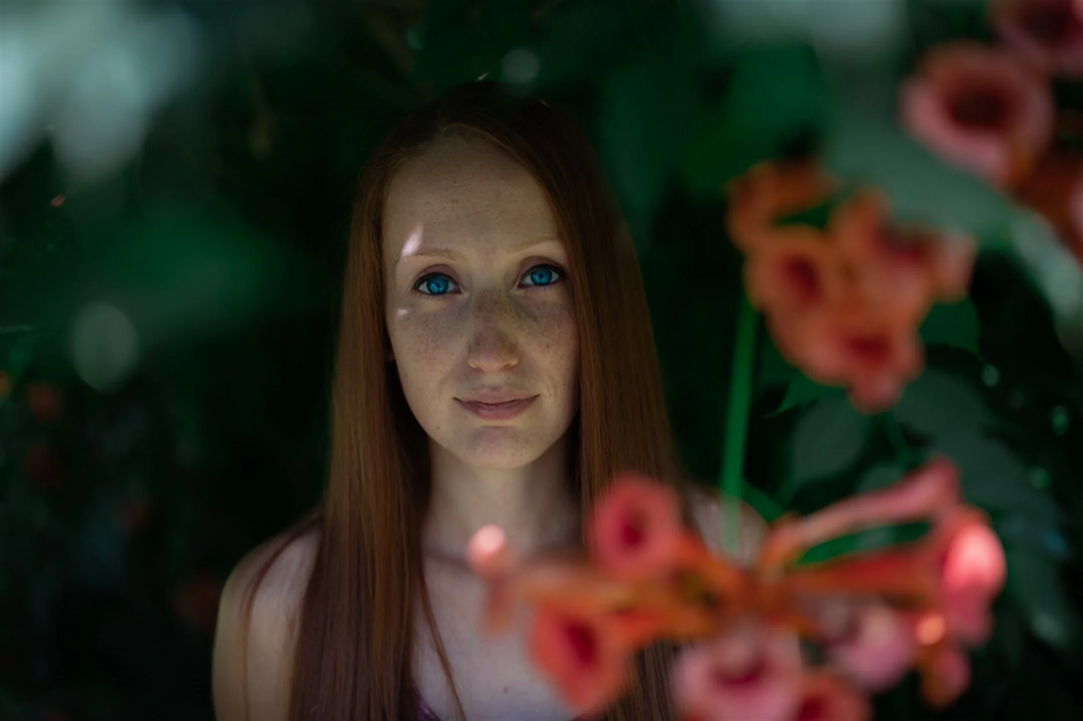 Portrait of a woman with long hair and freckles, surrounded by green foliage and blurred orange flowers in the foreground.