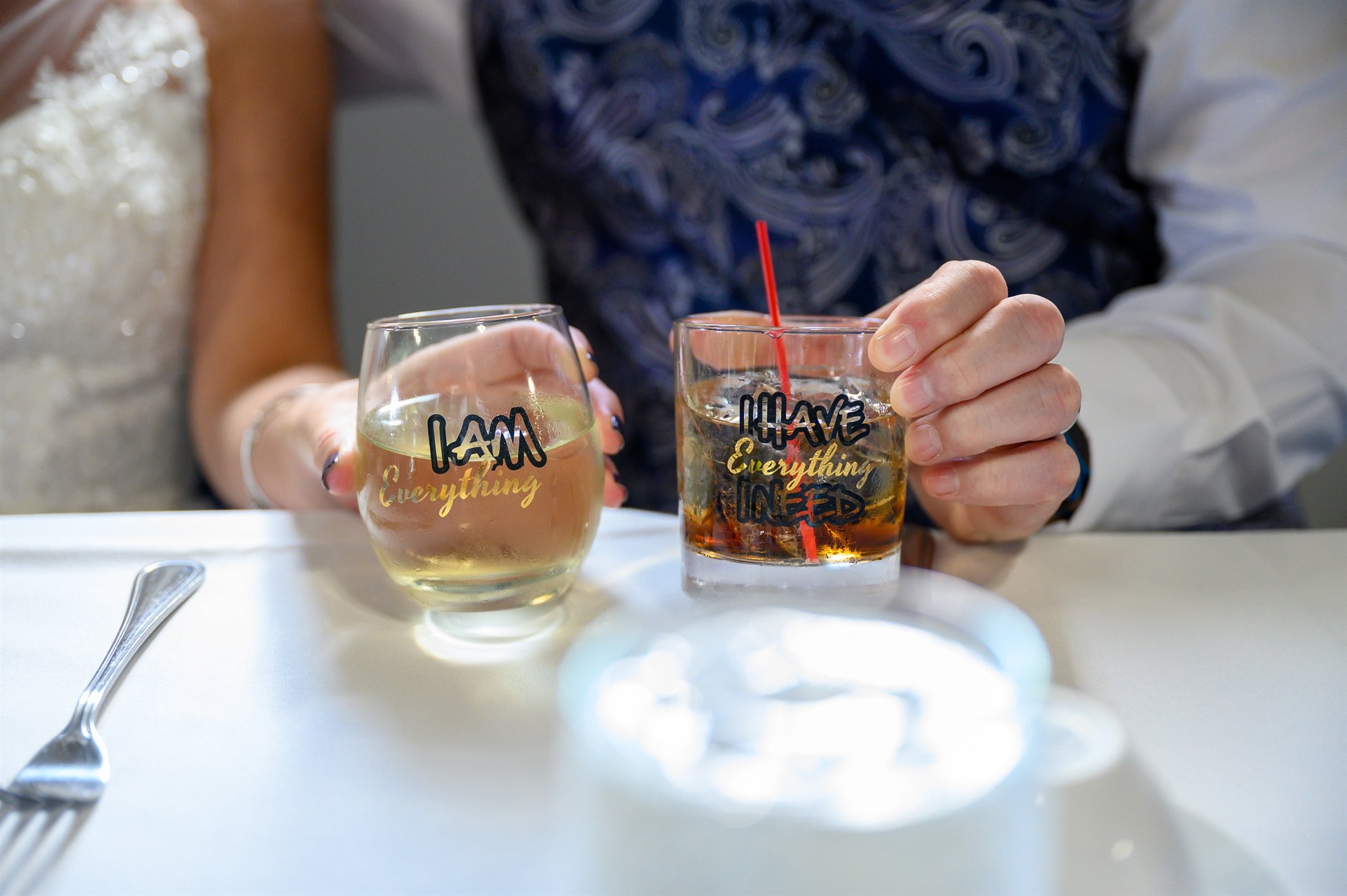 Two people holding glasses with different drinks, one with "I AM Everything" and the other with "I HAVE Everything I NEED" text, on a table with a fork.