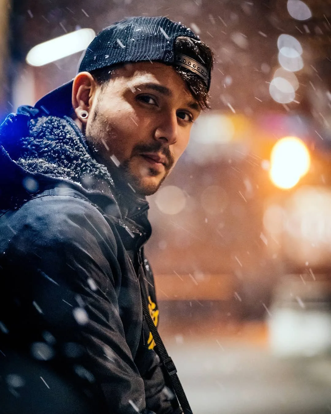 Man wearing a black baseball cap and jacket, standing in the snow at night, with bokeh lights in the background.