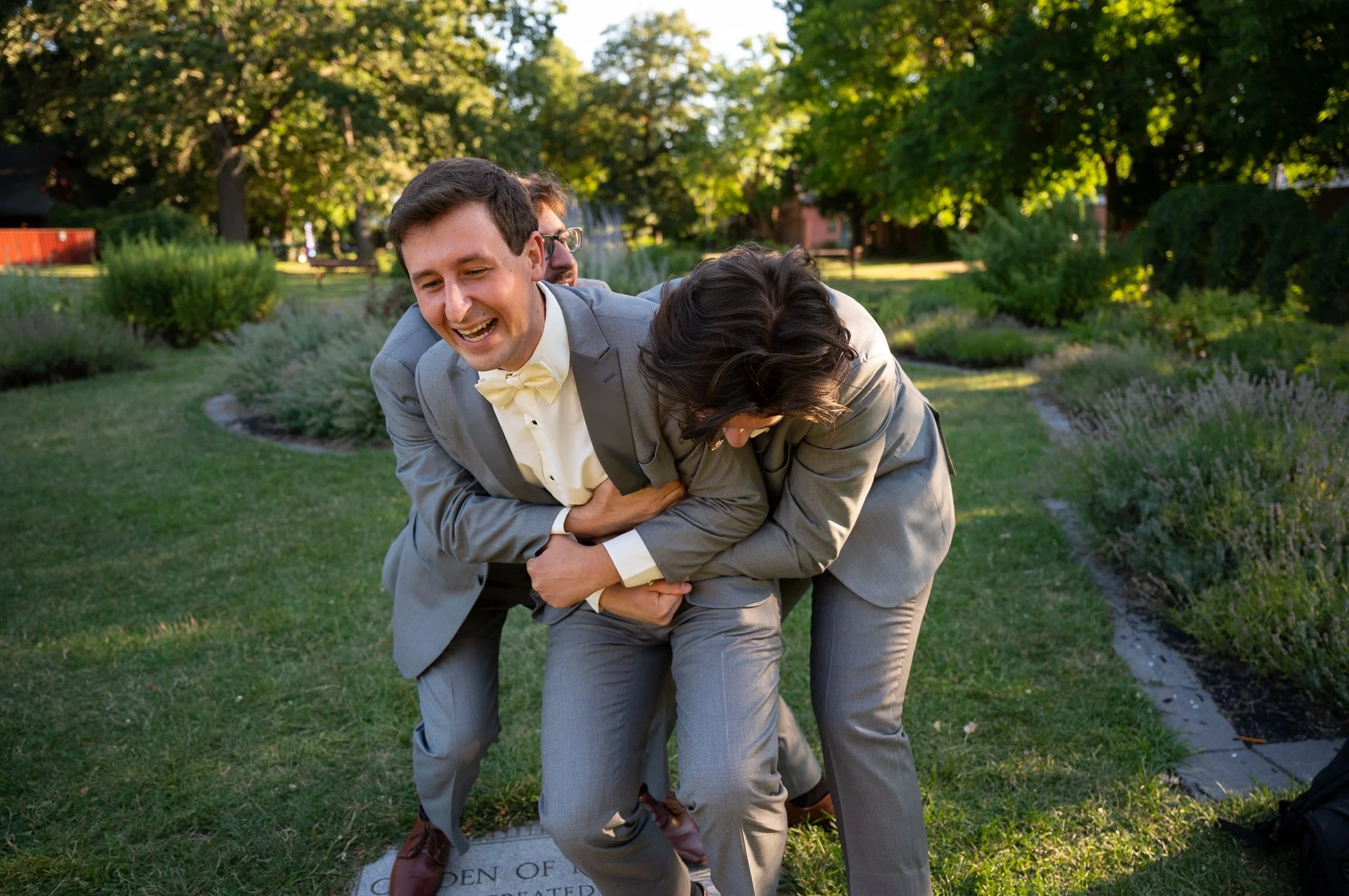 Two men in gray suits laughing and playfully hugging in a garden with trees and grass.