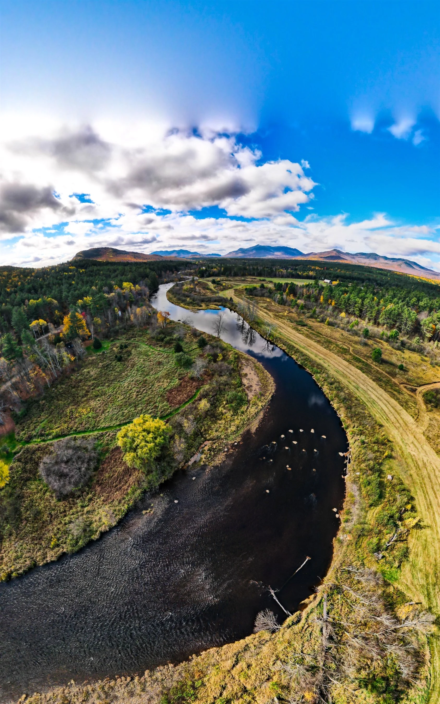 Aerial view of a winding river surrounded by lush greenery and forests, with mountains and a cloudy sky in the background.
