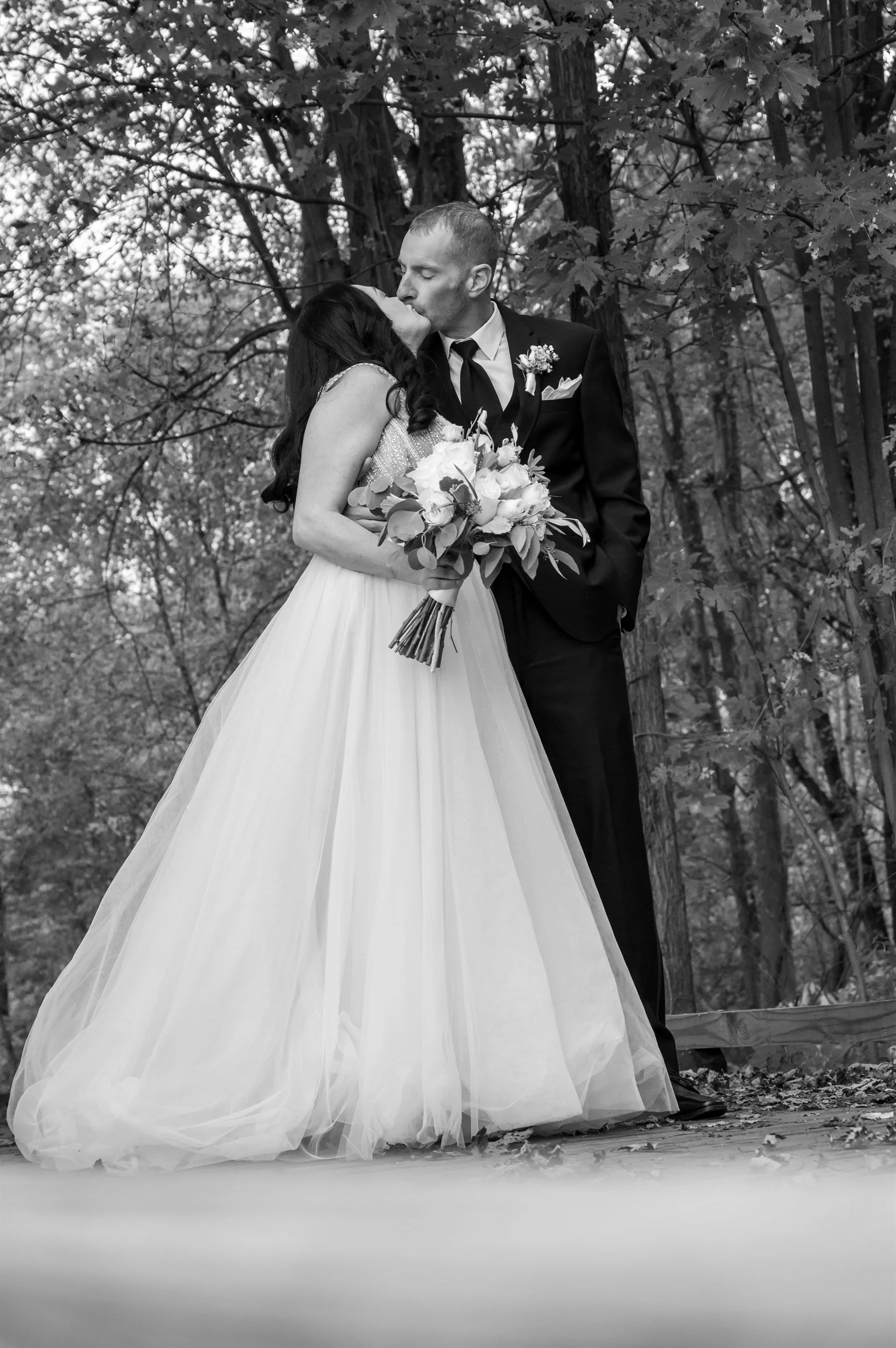 Black and white wedding photo of a bride in a gown and a groom in a suit kissing in a forest setting.