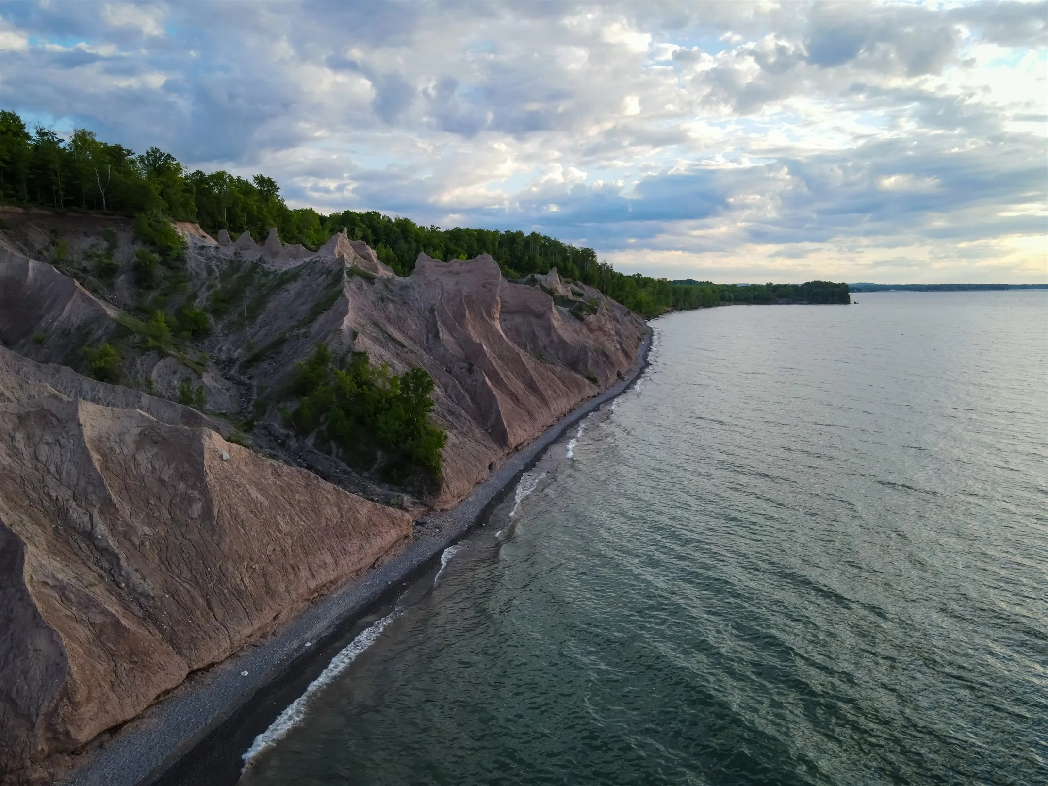 Eroded clay cliffs along a lake shoreline under a partly cloudy sky, with trees on the cliff edge.