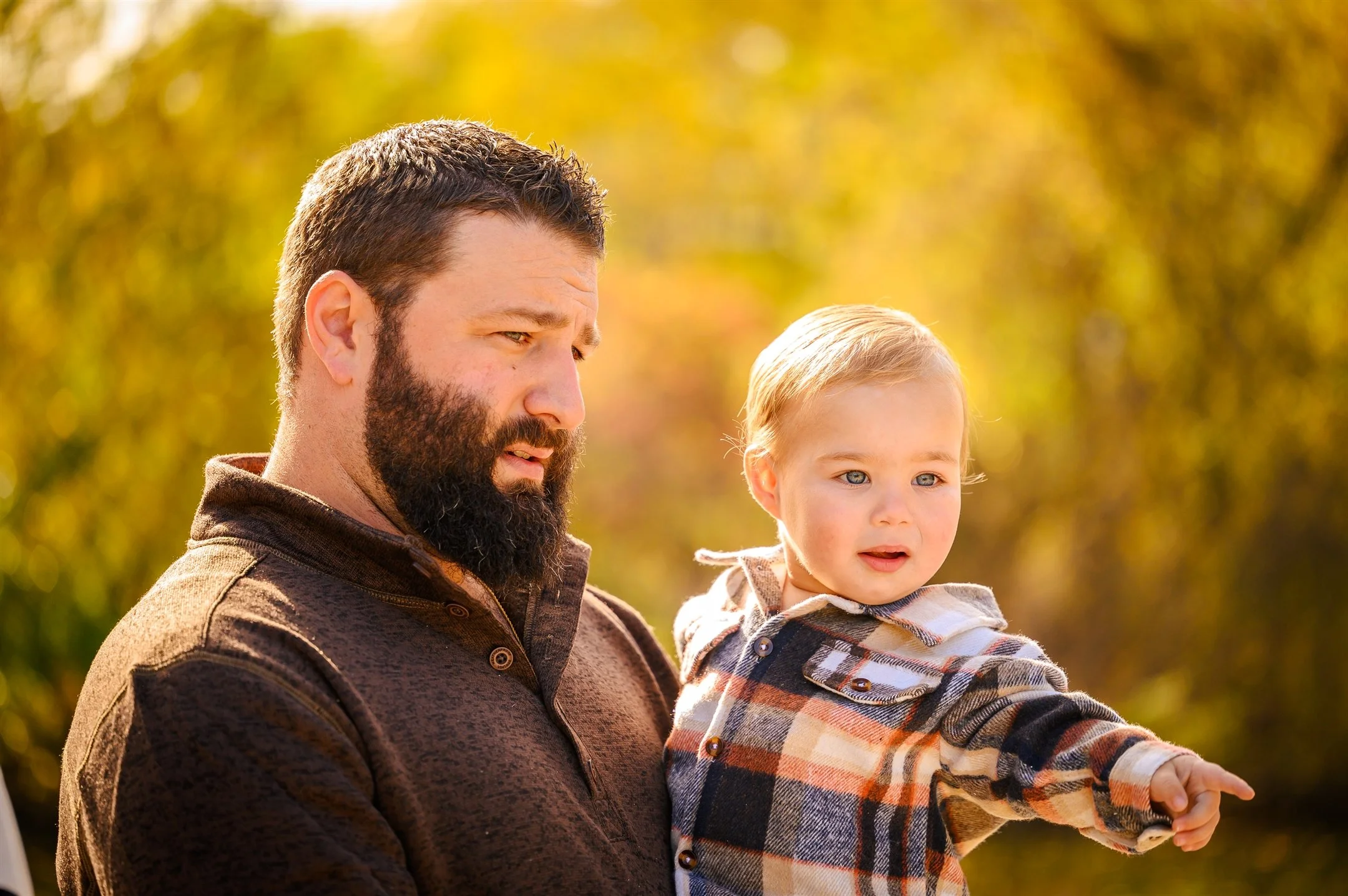 Man holding a young child, both looking in the same direction, with a blurred natural background.