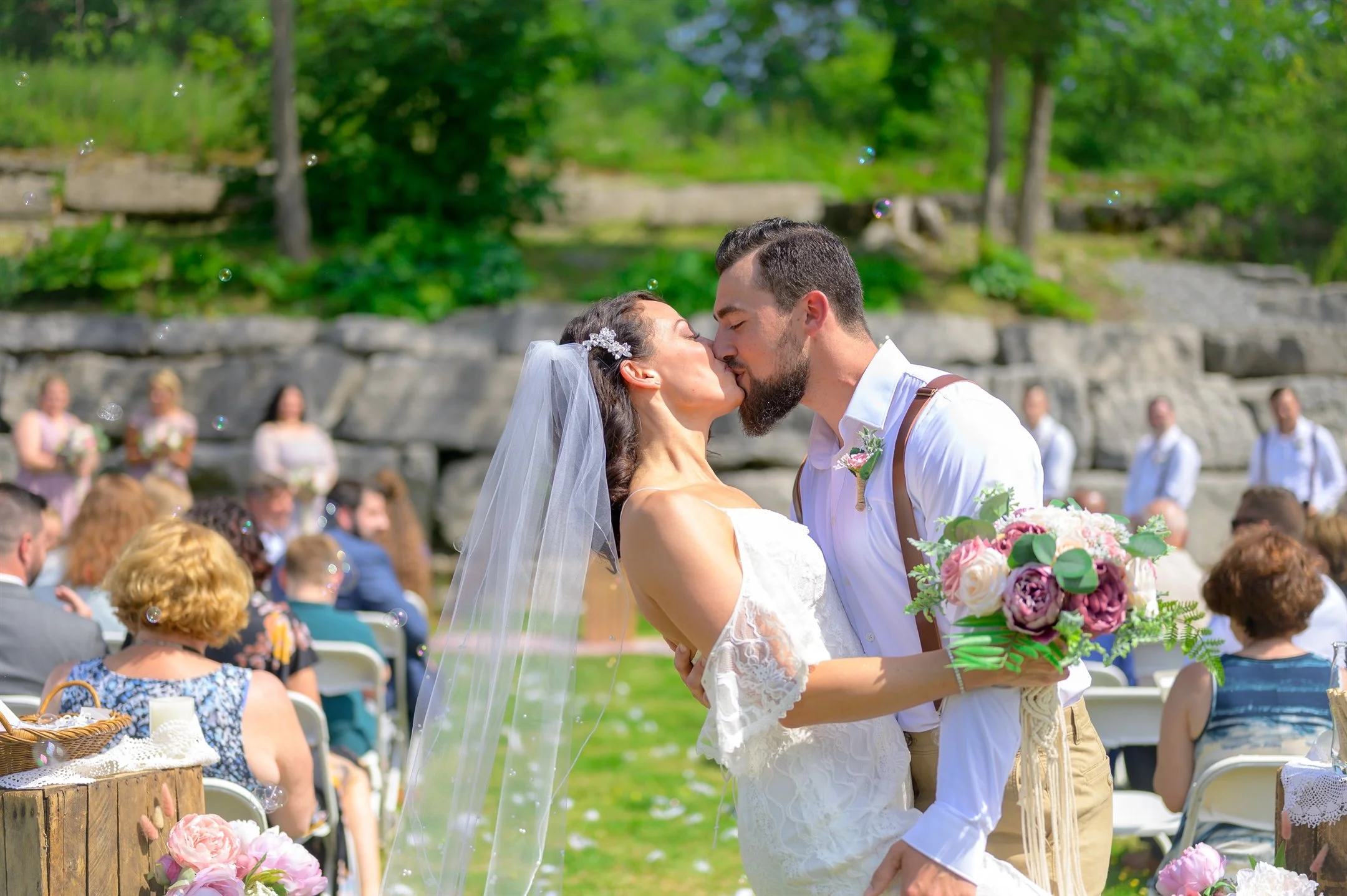 A bride and groom kissing at an outdoor wedding ceremony, surrounded by guests seated on white chairs, with greenery in the background.
