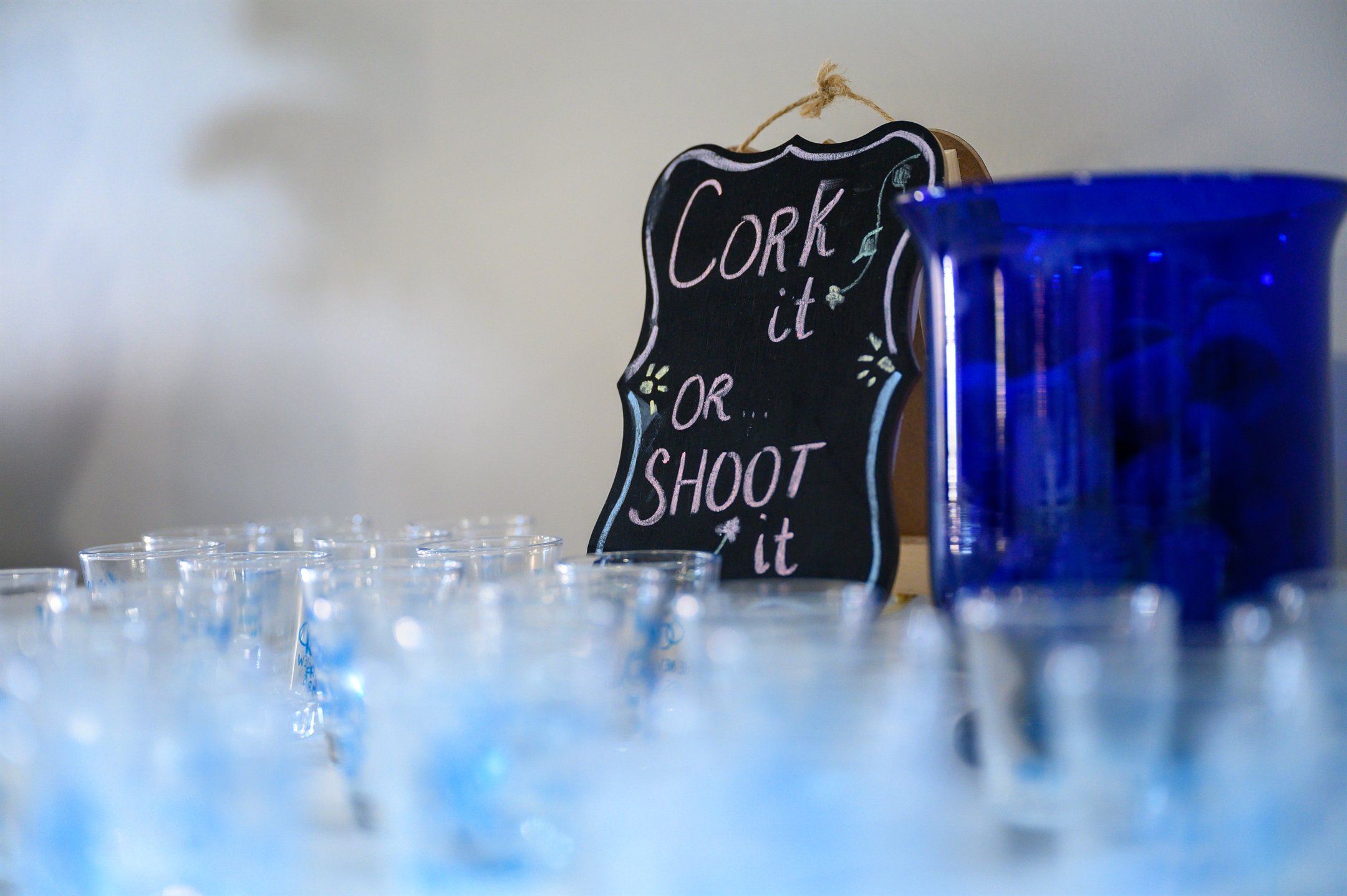 Shot glasses and blue pitcher with "Cork it or Shoot it" sign in background.
