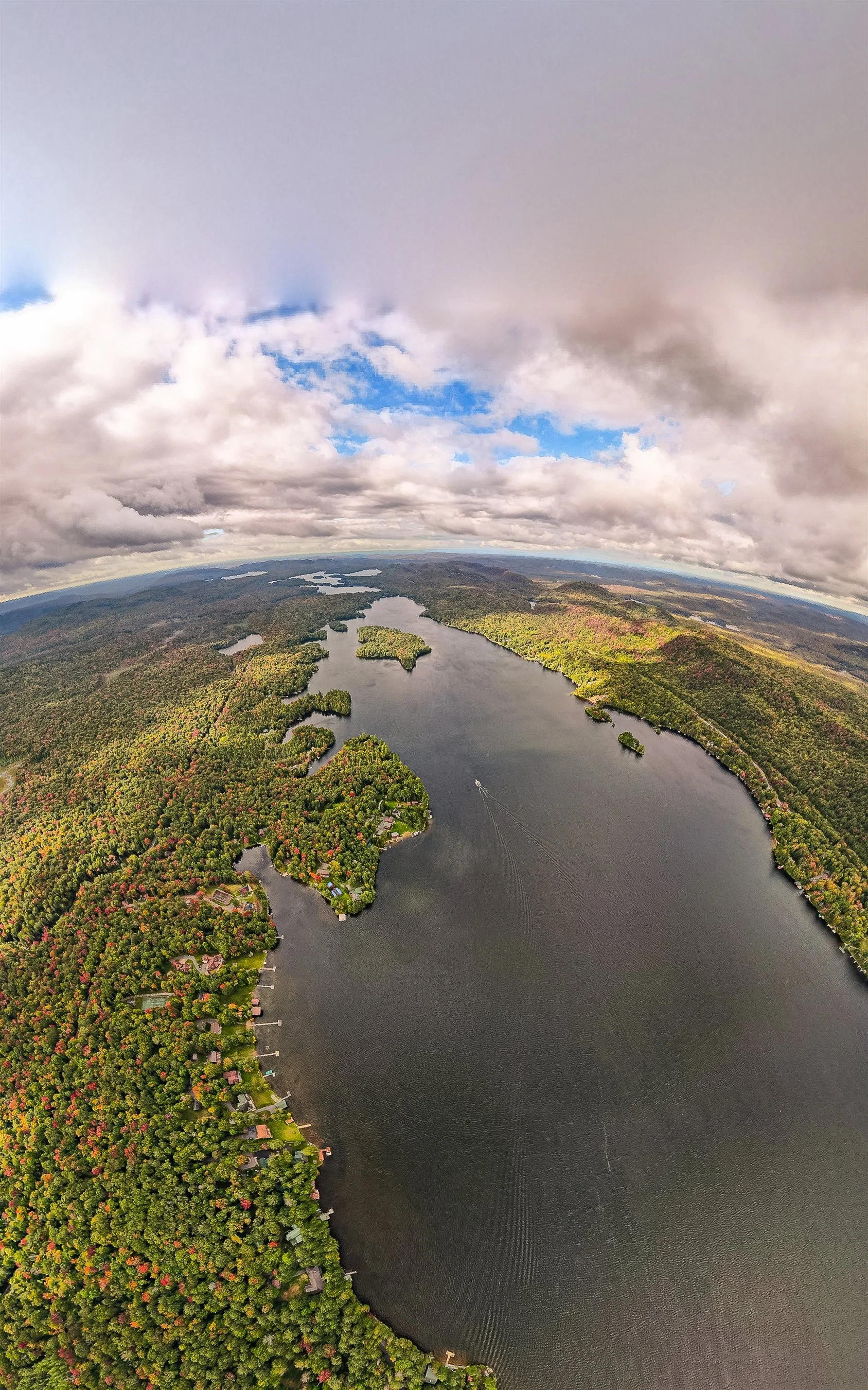 Aerial view of a large scenic lake surrounded by lush green forest with autumn foliage, under a partly cloudy sky.