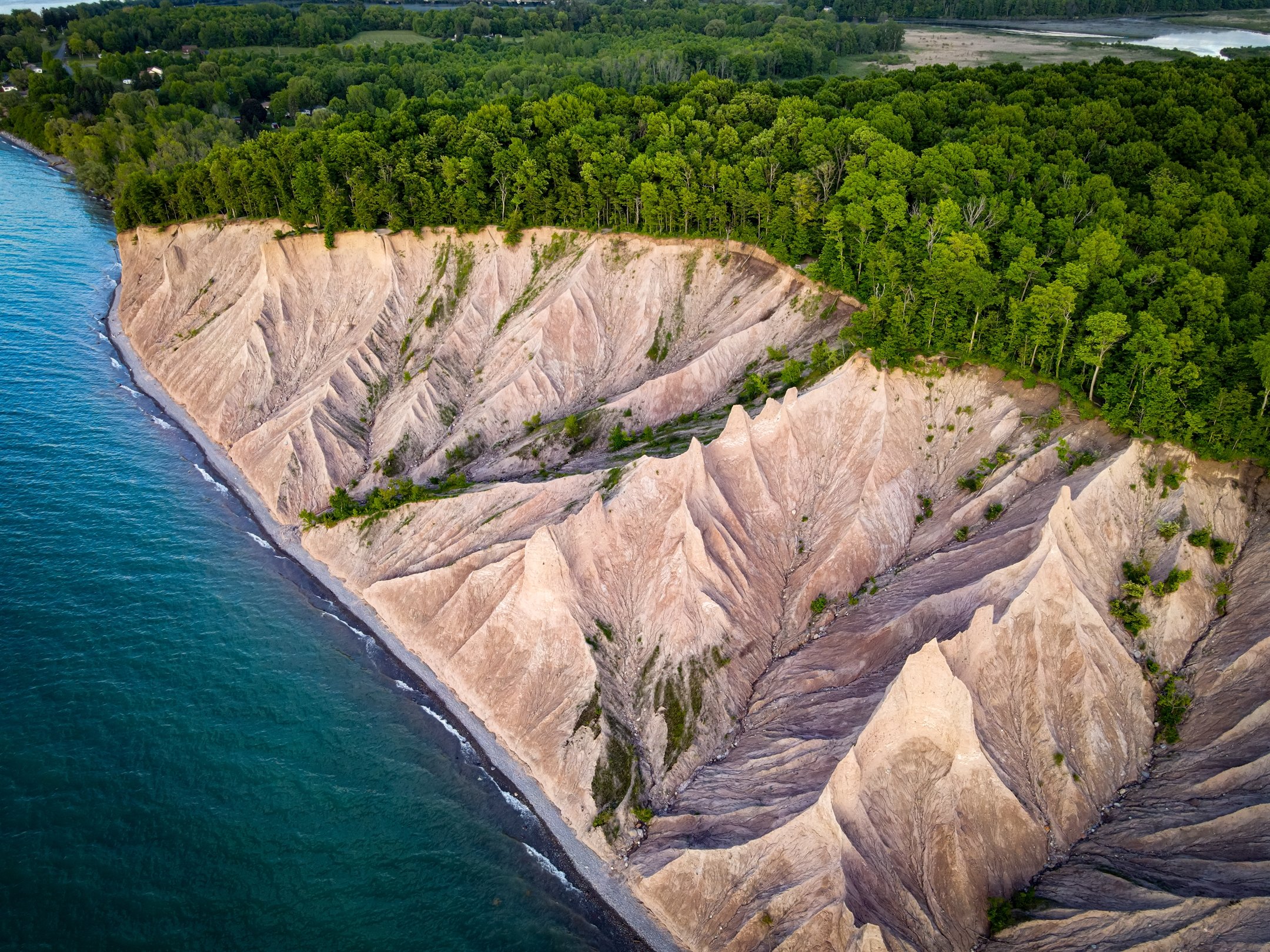 Aerial view of Chimney Bluffs State Park showing rugged sandstone cliffs, lush green forest, and a blue lake shoreline.