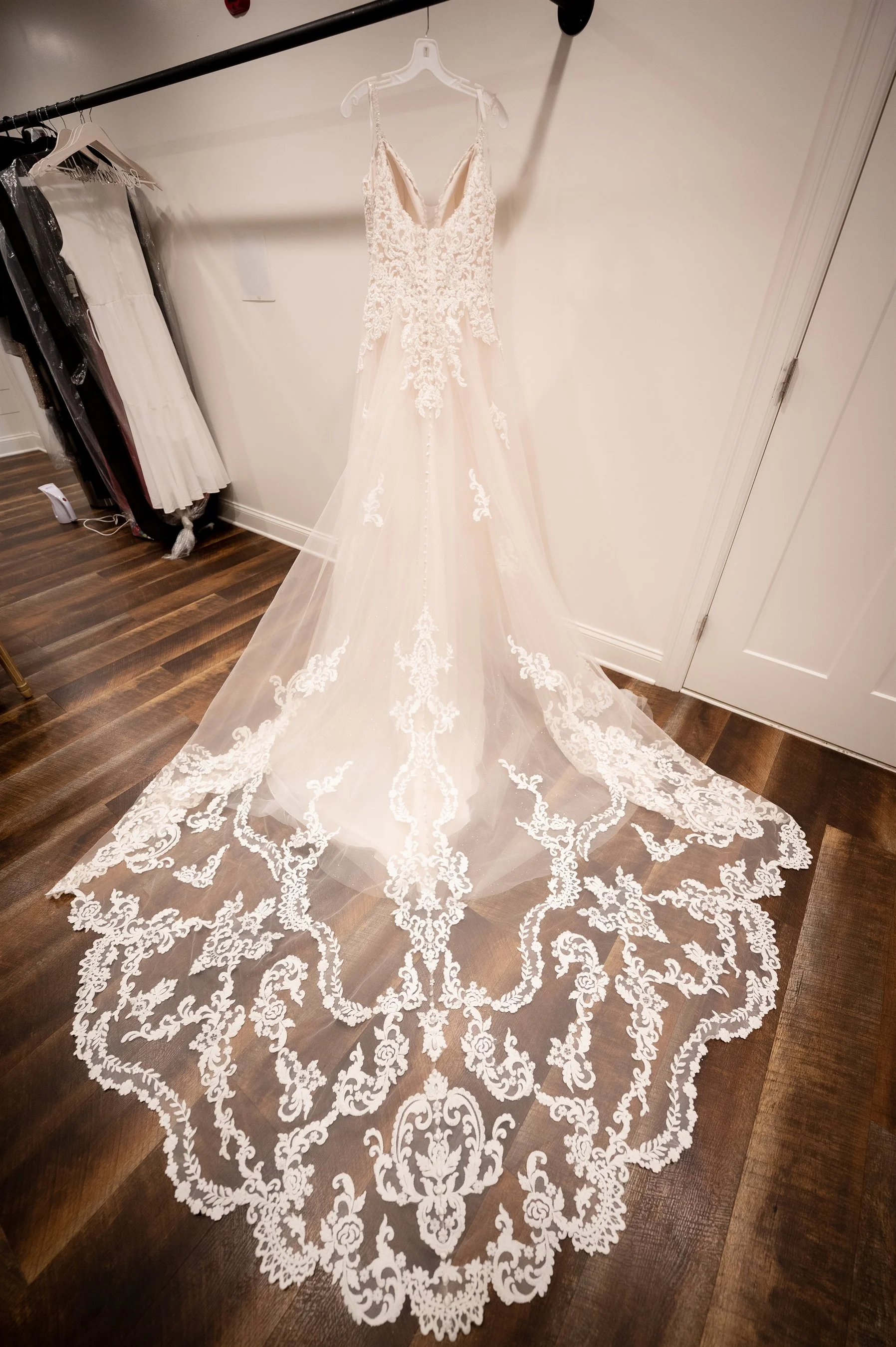 White lace wedding dress with intricate detailing on a hanger in a room with wooden floors.