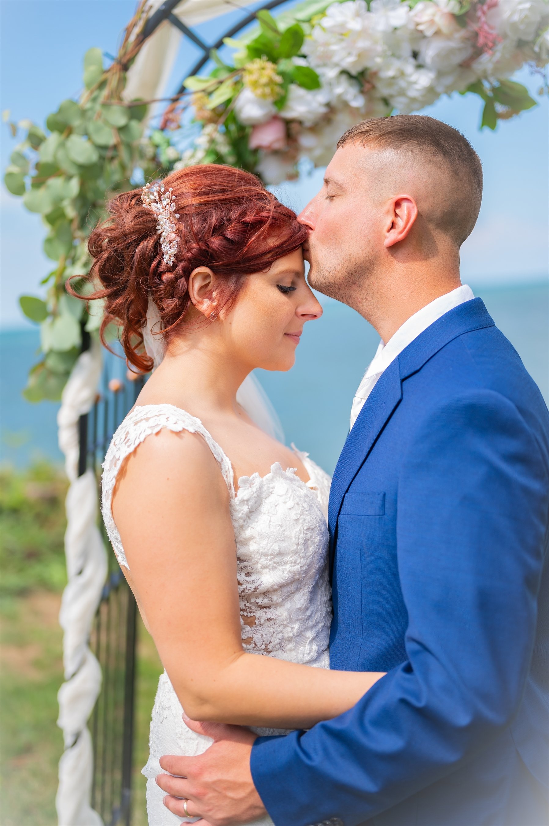 Wedding couple with groom kissing bride's forehead under floral arch by the sea.