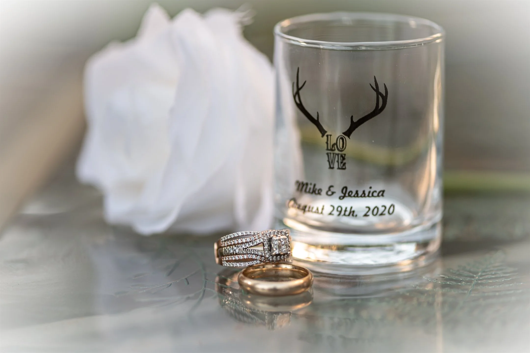 Close-up of wedding rings on a glass surface next to a white rose and a personalized glass with antler design and text 'LOVE' and wedding details.