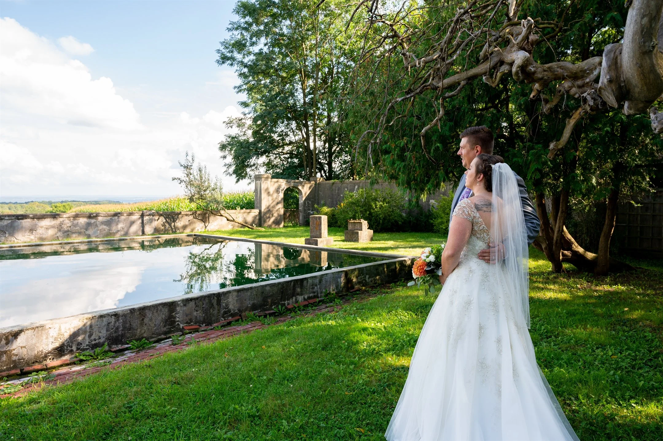 A couple in wedding attire standing near a reflective pool in a lush garden, surrounded by trees and a stone wall.