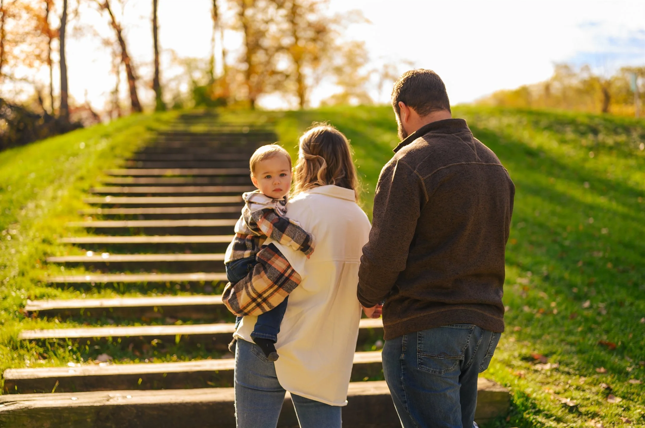A family of three standing outdoors in a park, with wooden steps and trees in the background; a man and woman facing away, holding a child who is looking at the camera.