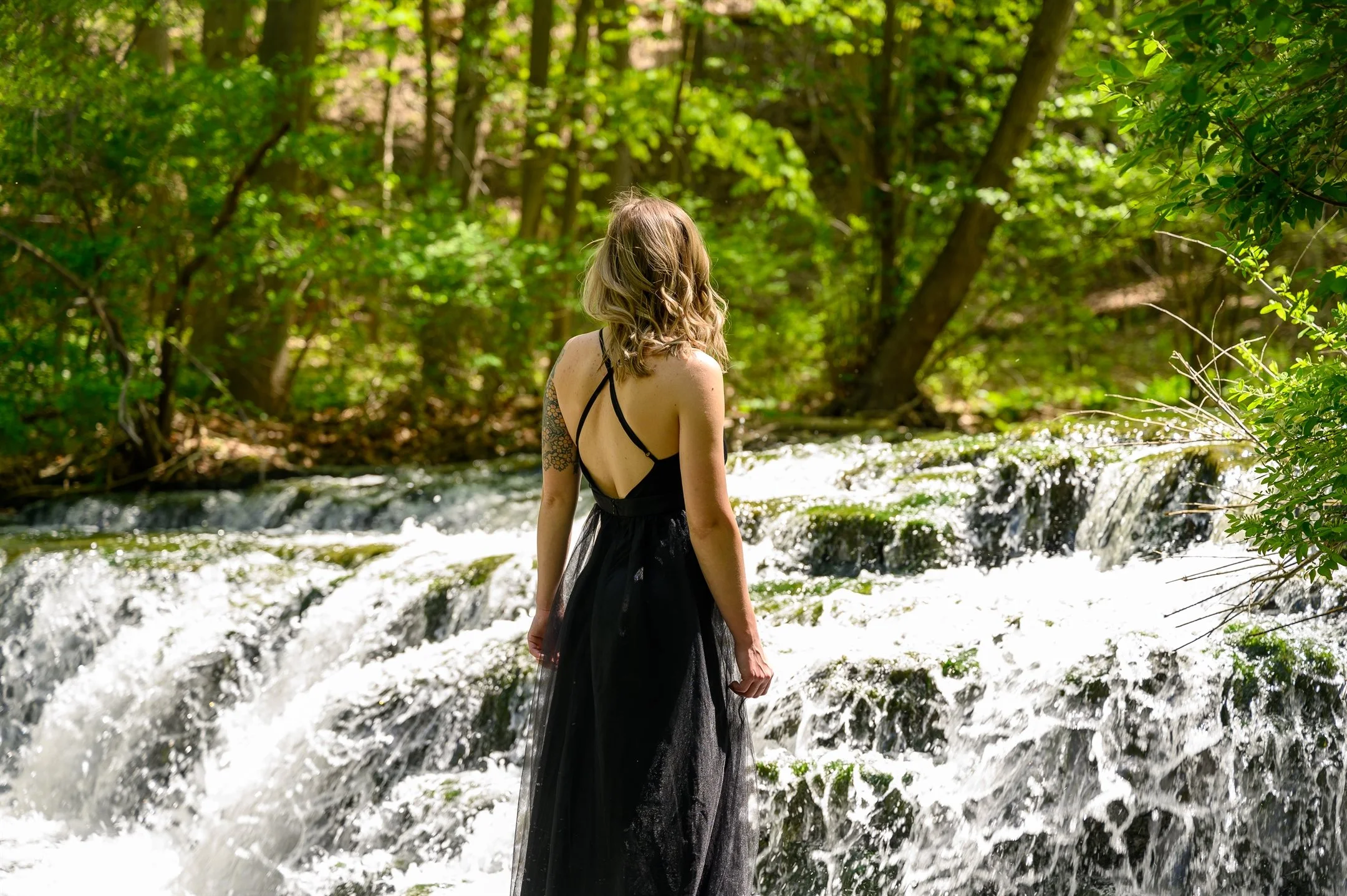 Woman in black dress standing near a small waterfall in a forest