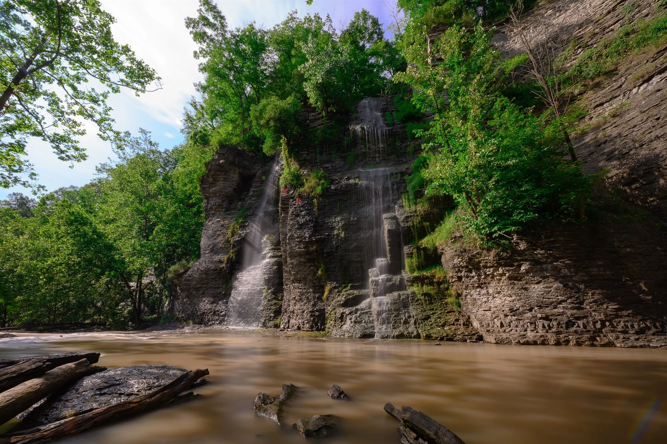 Serene waterfall cascading over rocky cliffs surrounded by lush green foliage and trees with a tranquil pool below.