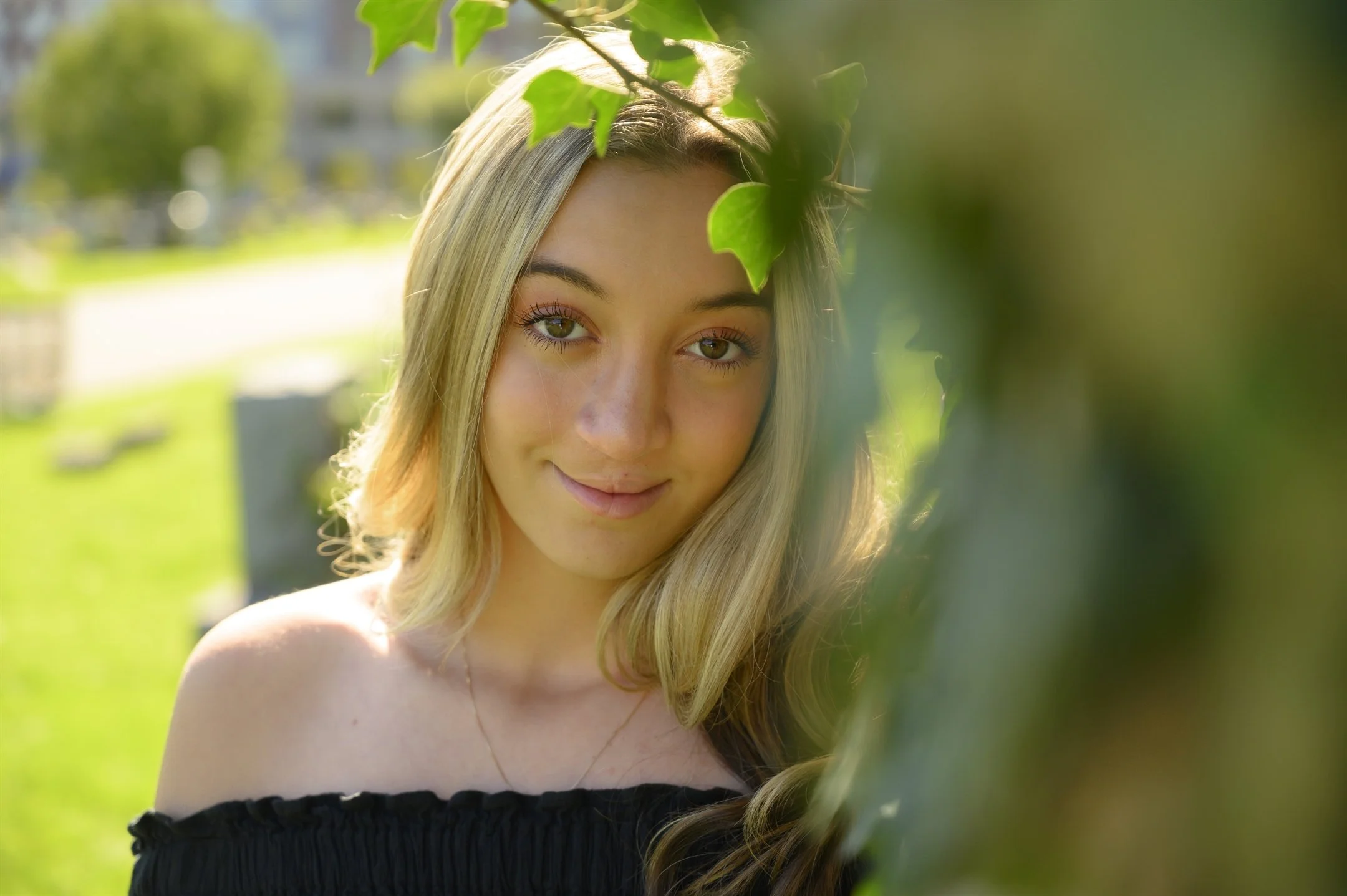 Young woman with long blonde hair, standing outdoors near leafy branches, wearing a black off-shoulder top, smiling gently.