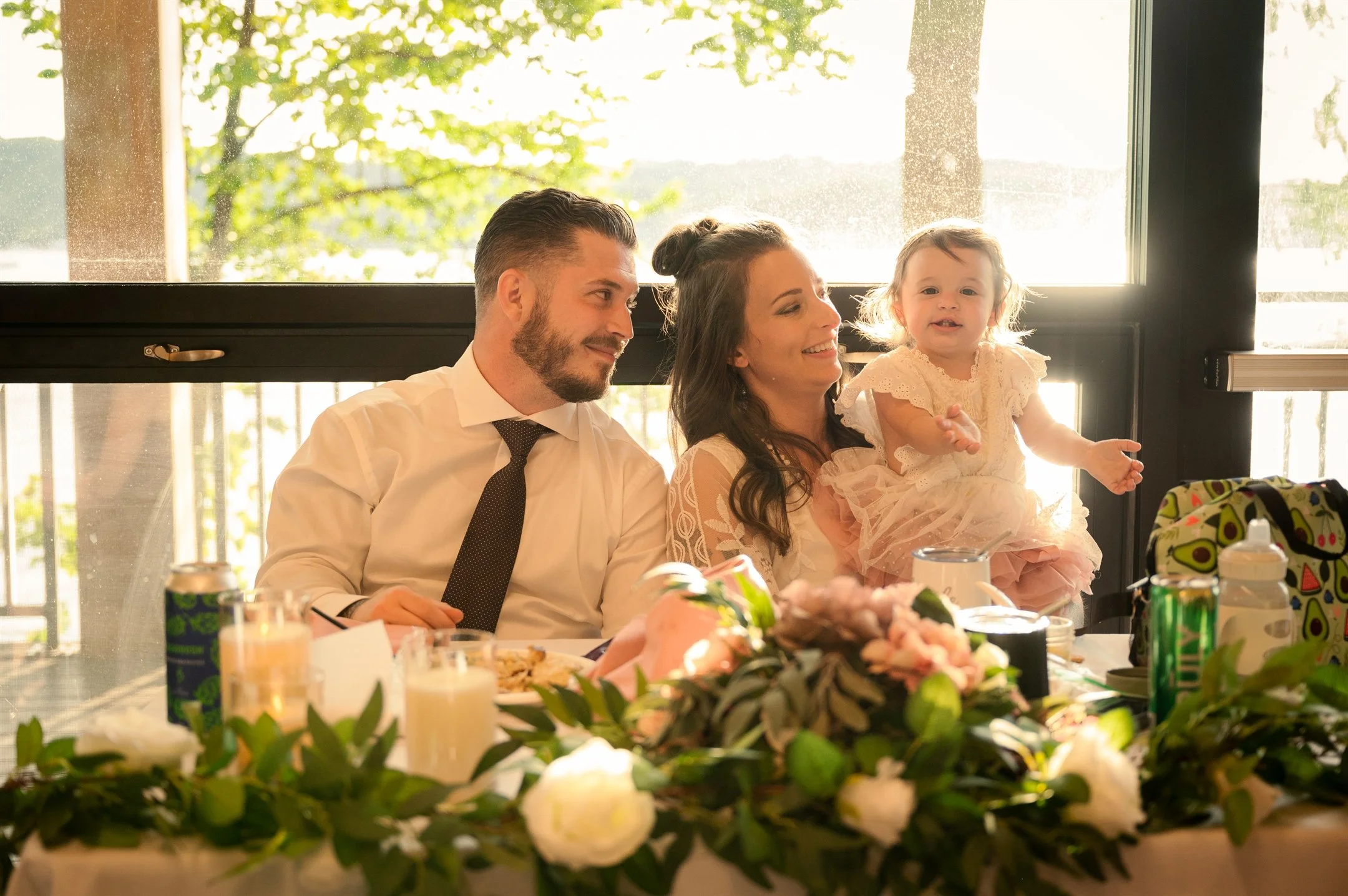 A family sitting at a decorated table, smiling. The woman is holding a toddler wearing a dress, with sunlight streaming through a large window behind them. Green foliage and flowers adorn the table.