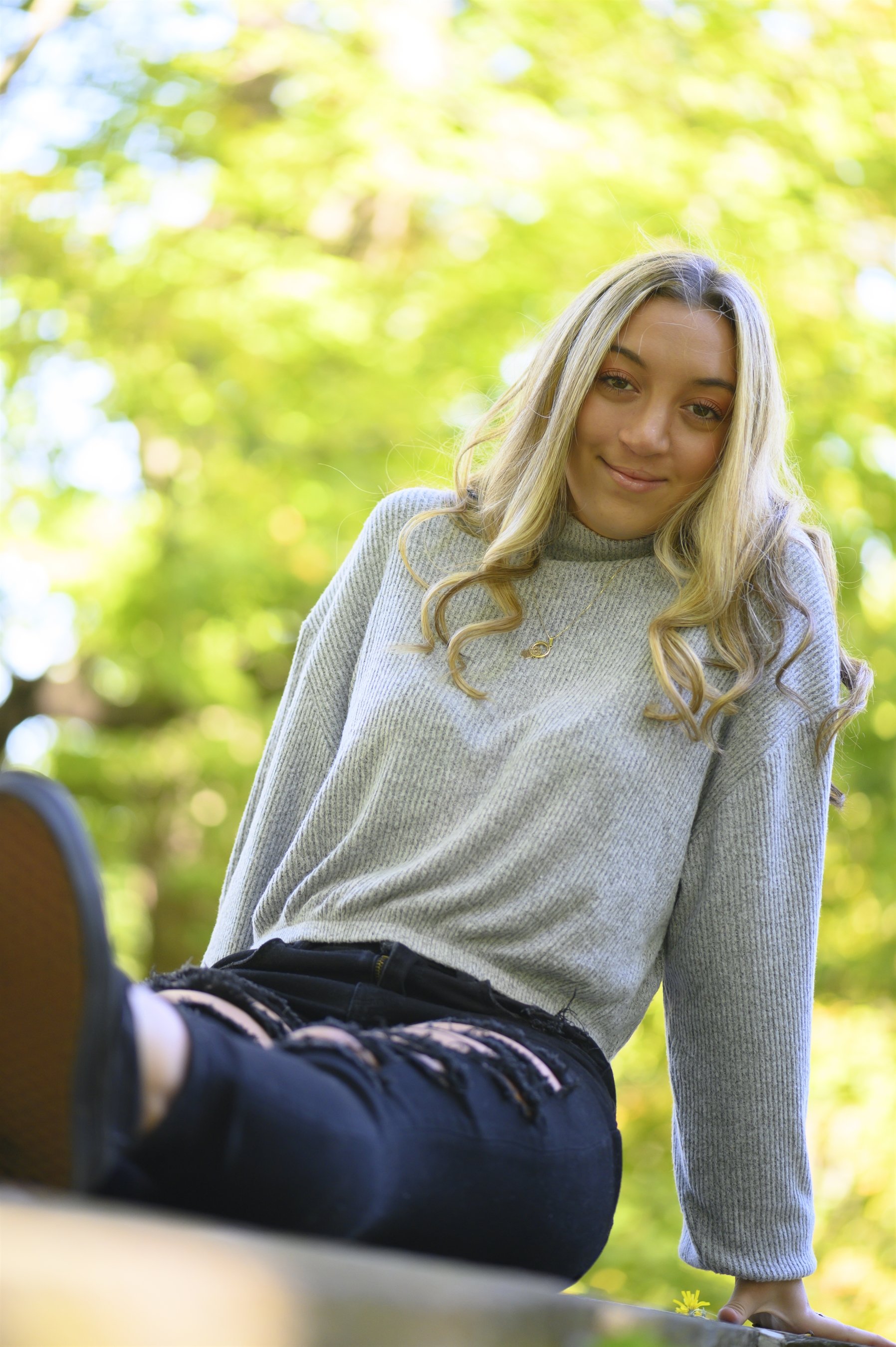 Woman with blonde hair in a gray sweater and ripped jeans sitting outdoors with trees in the background.