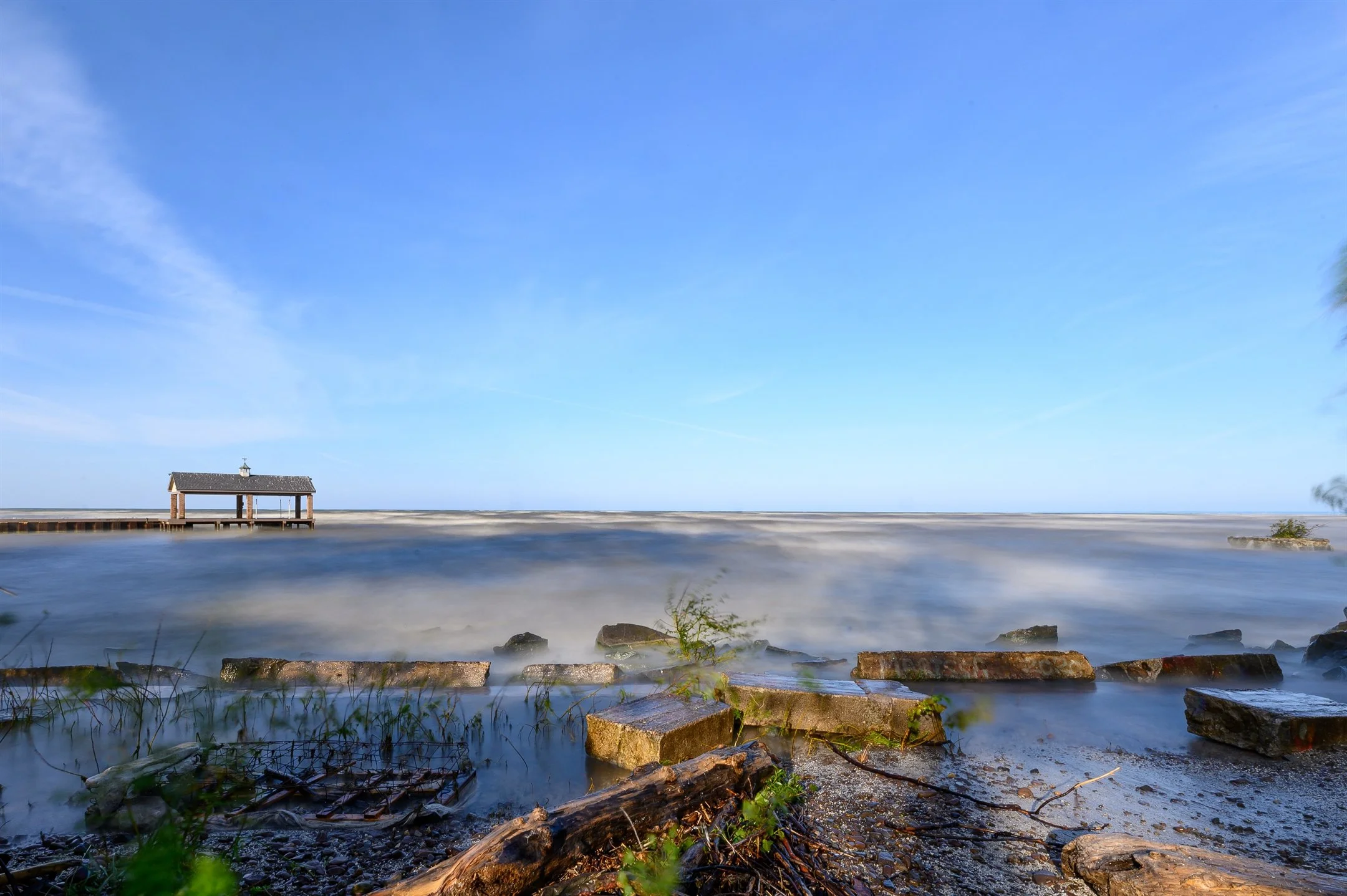 Lakeside view with a small wooden dock over water, cloudy blue sky, and rocky shoreline in the foreground.