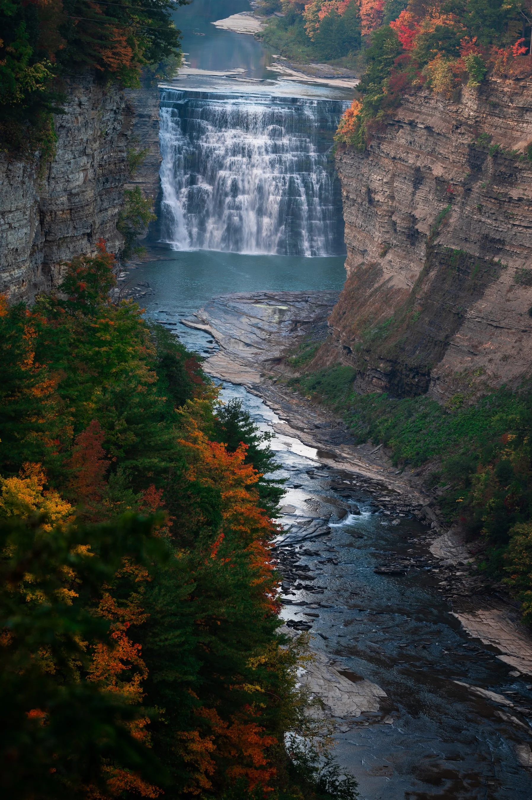 Scenic waterfall cascading between cliffs with colorful autumn trees, seen from above, and a river flowing below.
