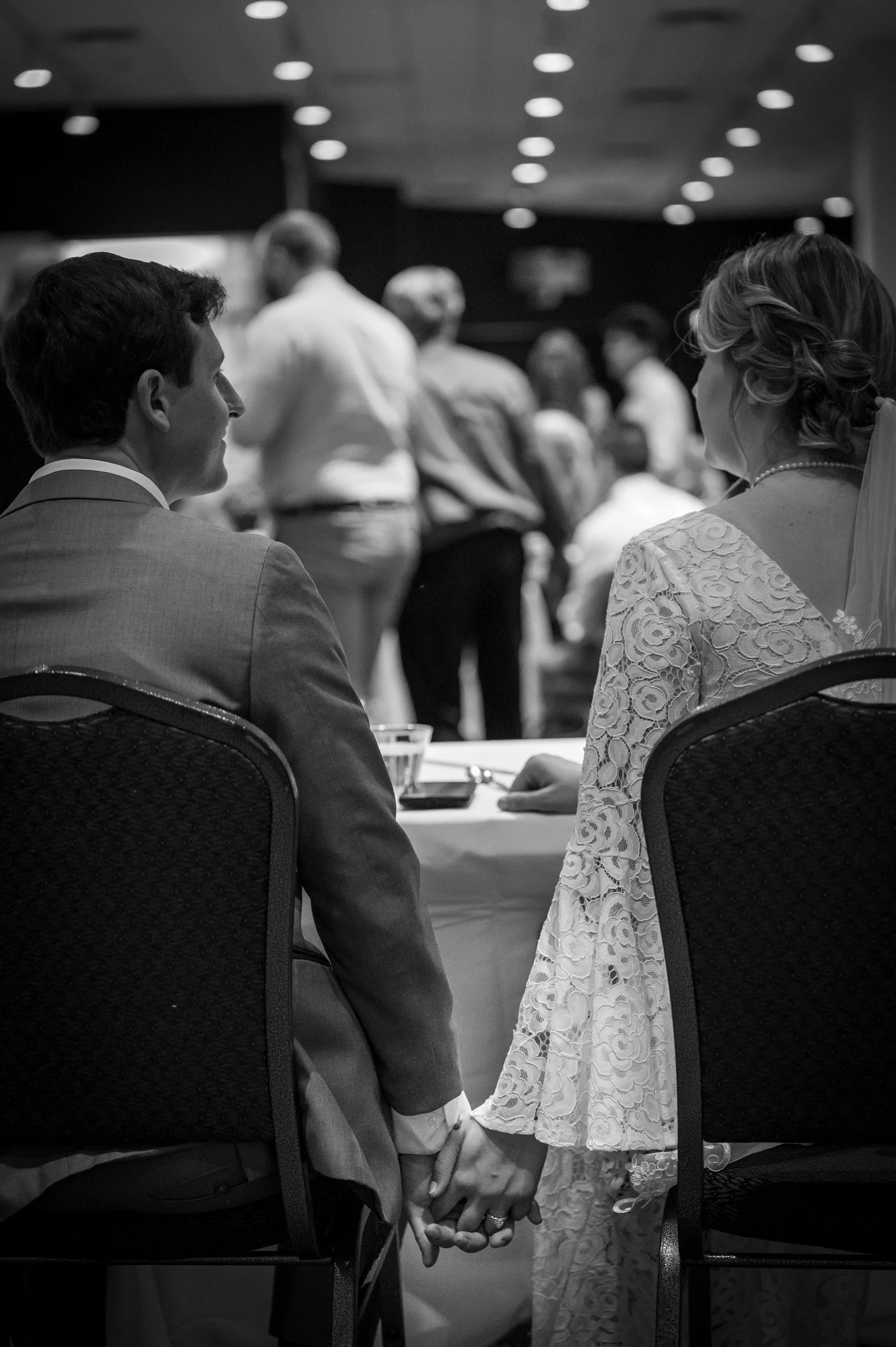Black and white photo of a bride and groom holding hands while seated at a table during a wedding reception.
