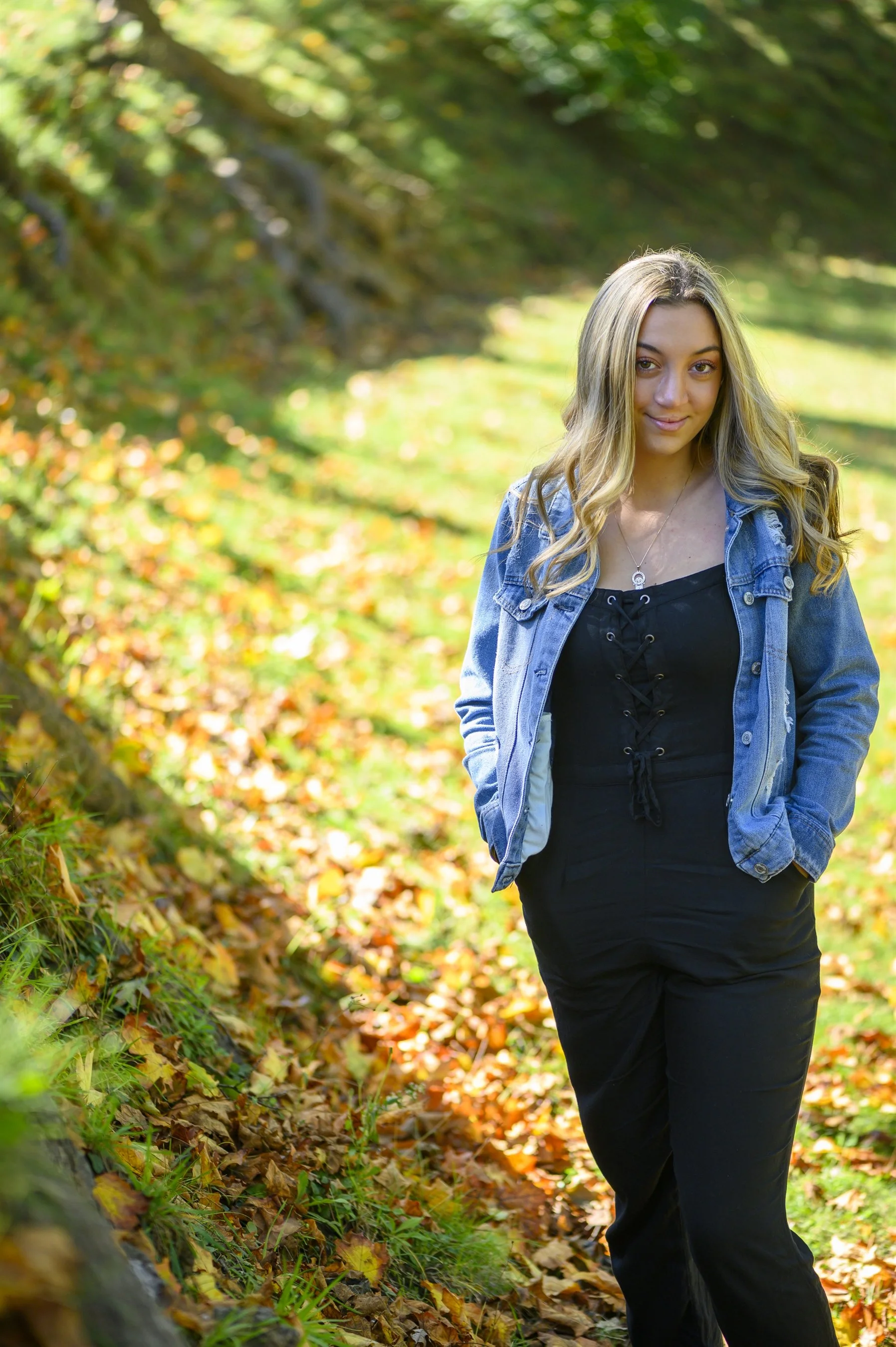 Woman in denim jacket and black jumpsuit standing outdoors on a grassy slope with autumn leaves.