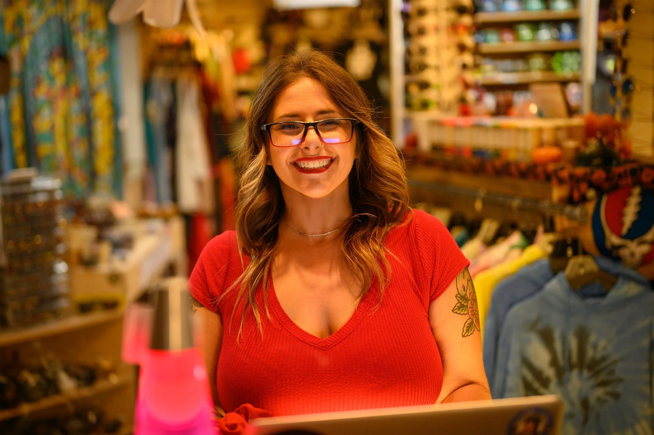Woman with glasses and tattoos smiling in clothing store, wearing a red shirt.