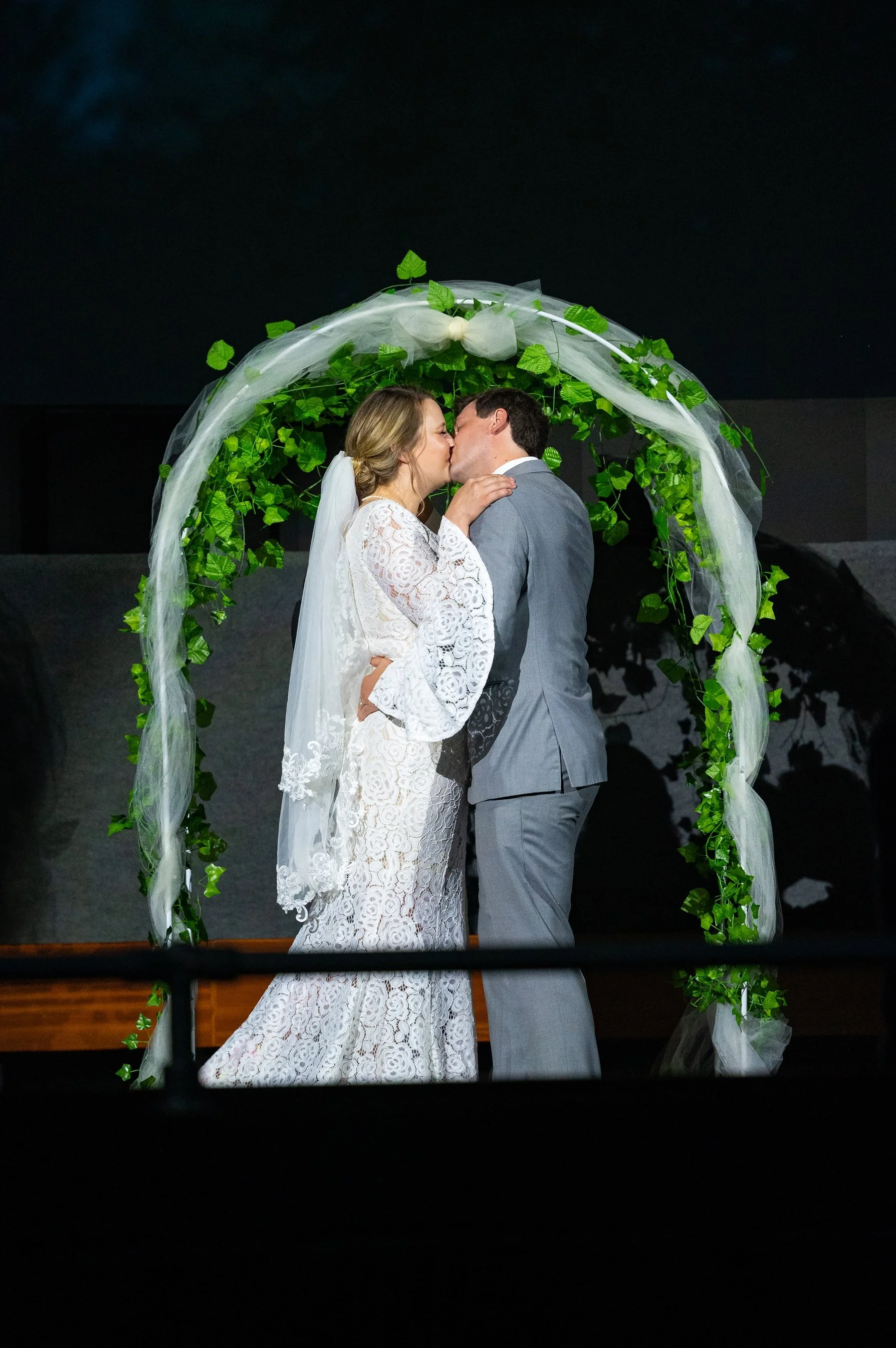 A bride and groom kissing under a wedding arch decorated with greenery and white fabric.