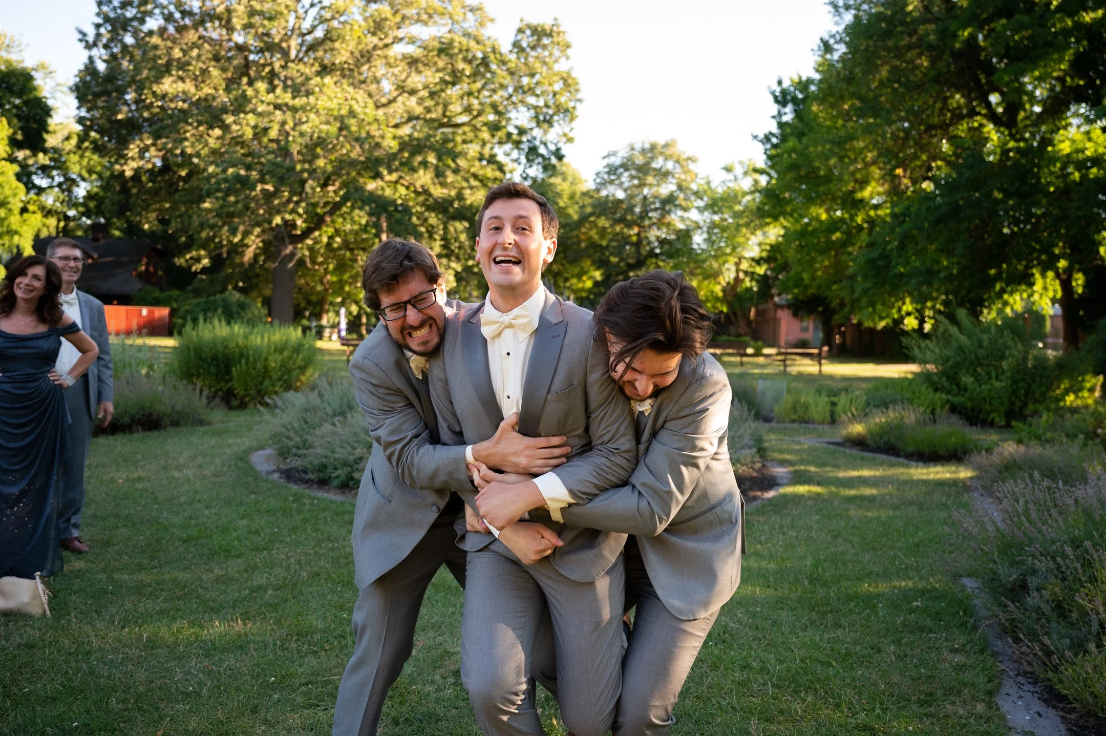 Three men in gray suits laughing and hugging in a garden, with two people in the background watching.