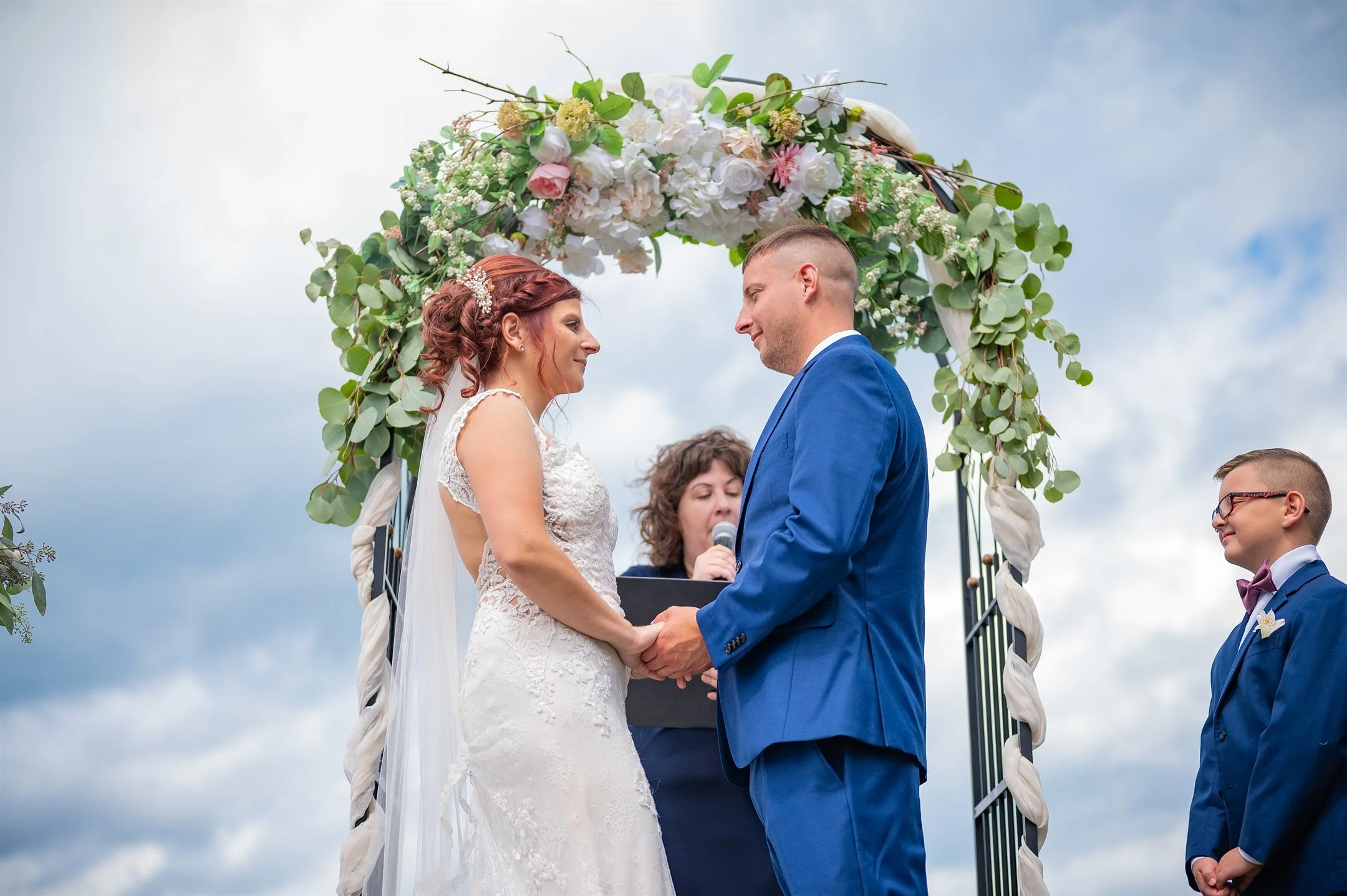 A bride and groom hold hands during an outdoor wedding ceremony under a floral arch, with a young boy standing beside them.