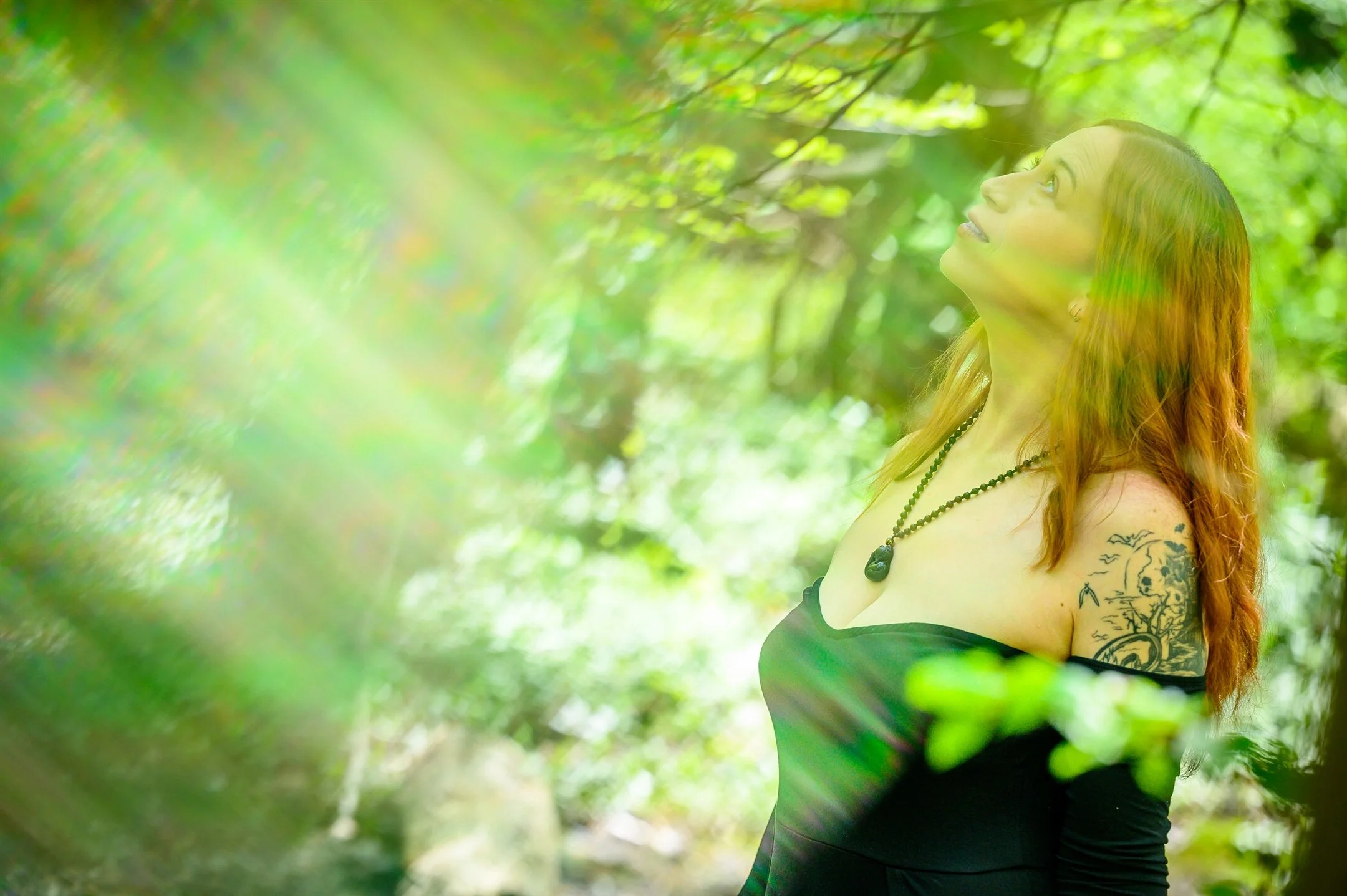 Woman with red hair in a black dress looking up in a forest, sunlight filtering through leaves, tattoo on arm, wearing a necklace.