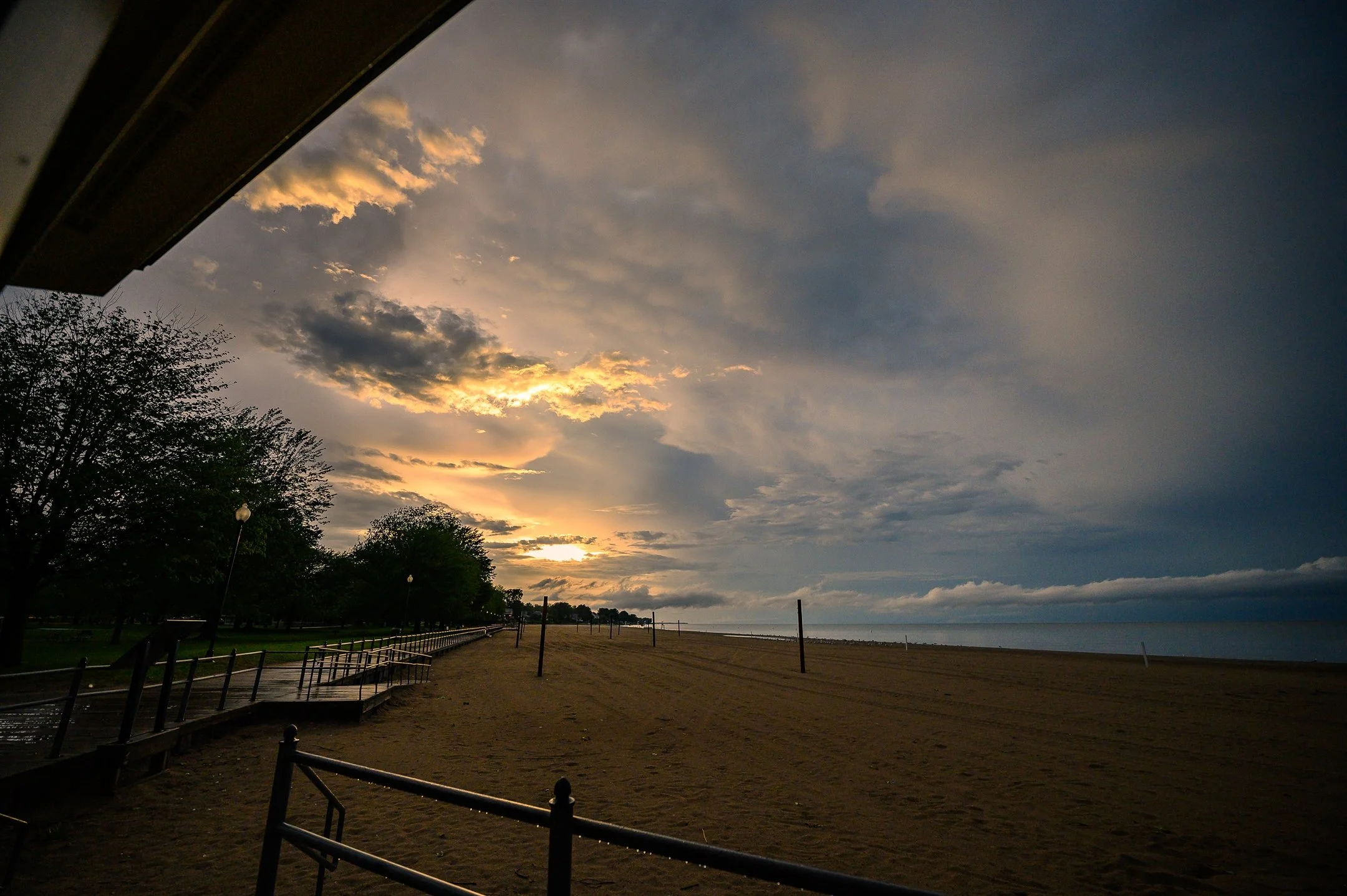 Beach scene at sunset with dramatic sky, empty sandy beach, railings and trees in the foreground, and ocean horizon in the background.