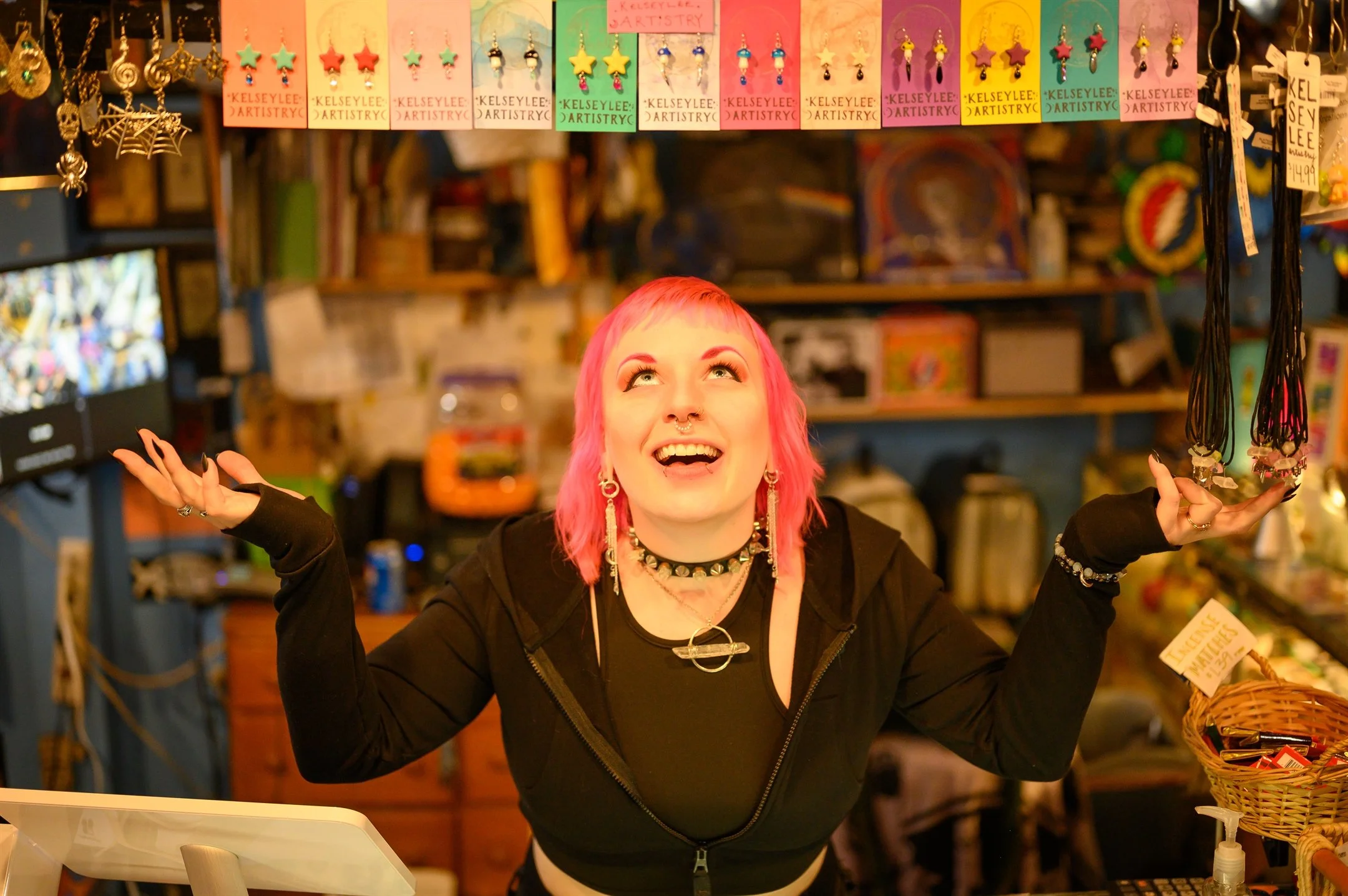 Person with pink hair and black clothing smiling in a colorful shop with hanging earrings and necklaces.