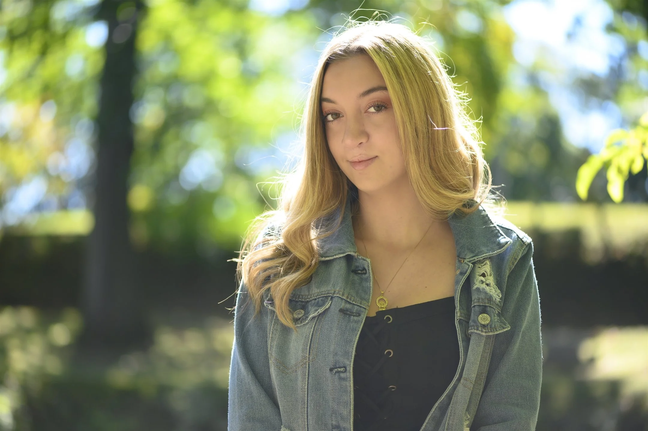 Young woman in denim jacket, outdoors with blurred trees in the background.