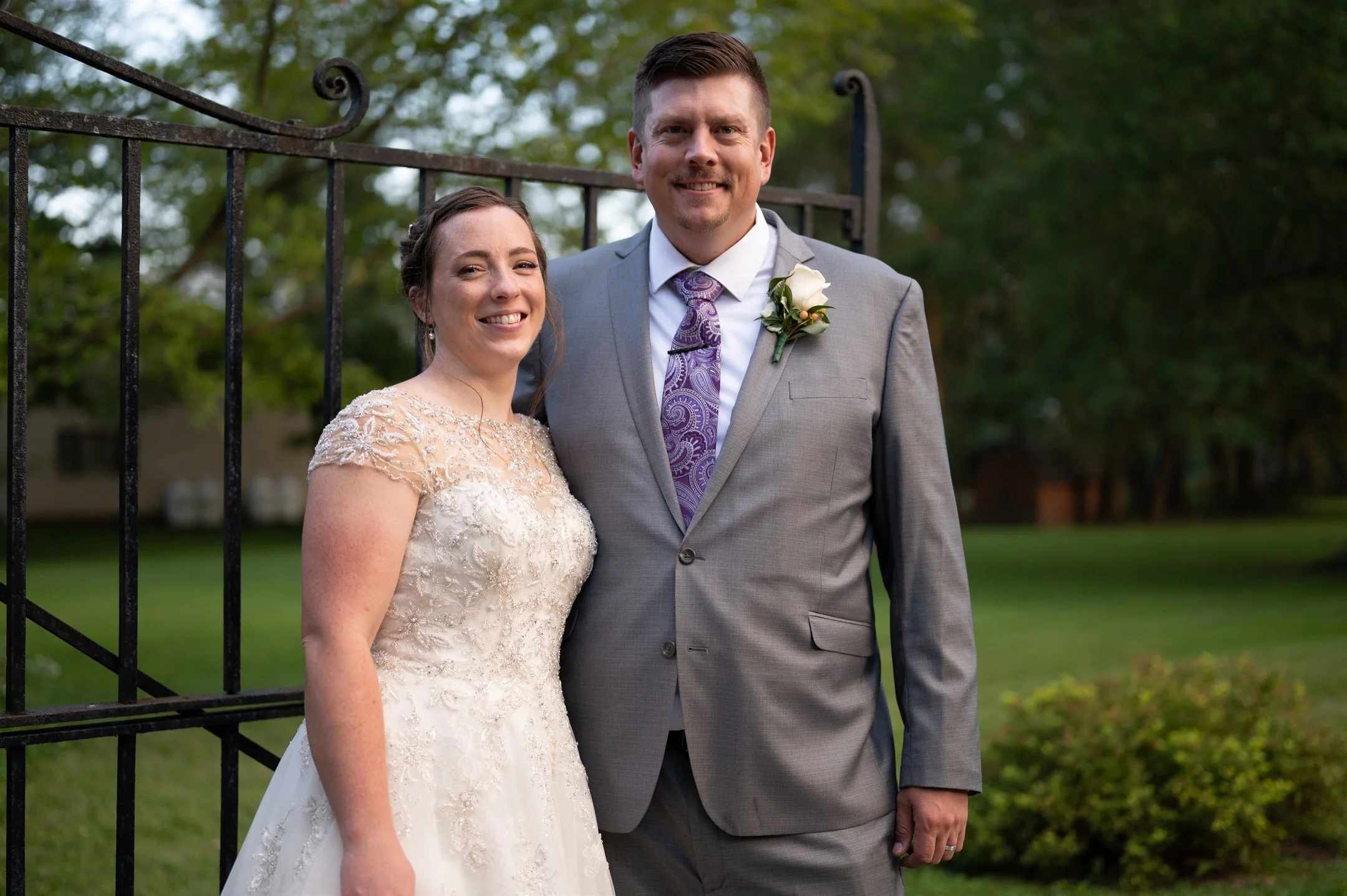 A couple dressed in formal attire smiling in front of a decorative black iron gate; the woman in a white wedding dress, the man in a gray suit with a purple tie and boutonniere.