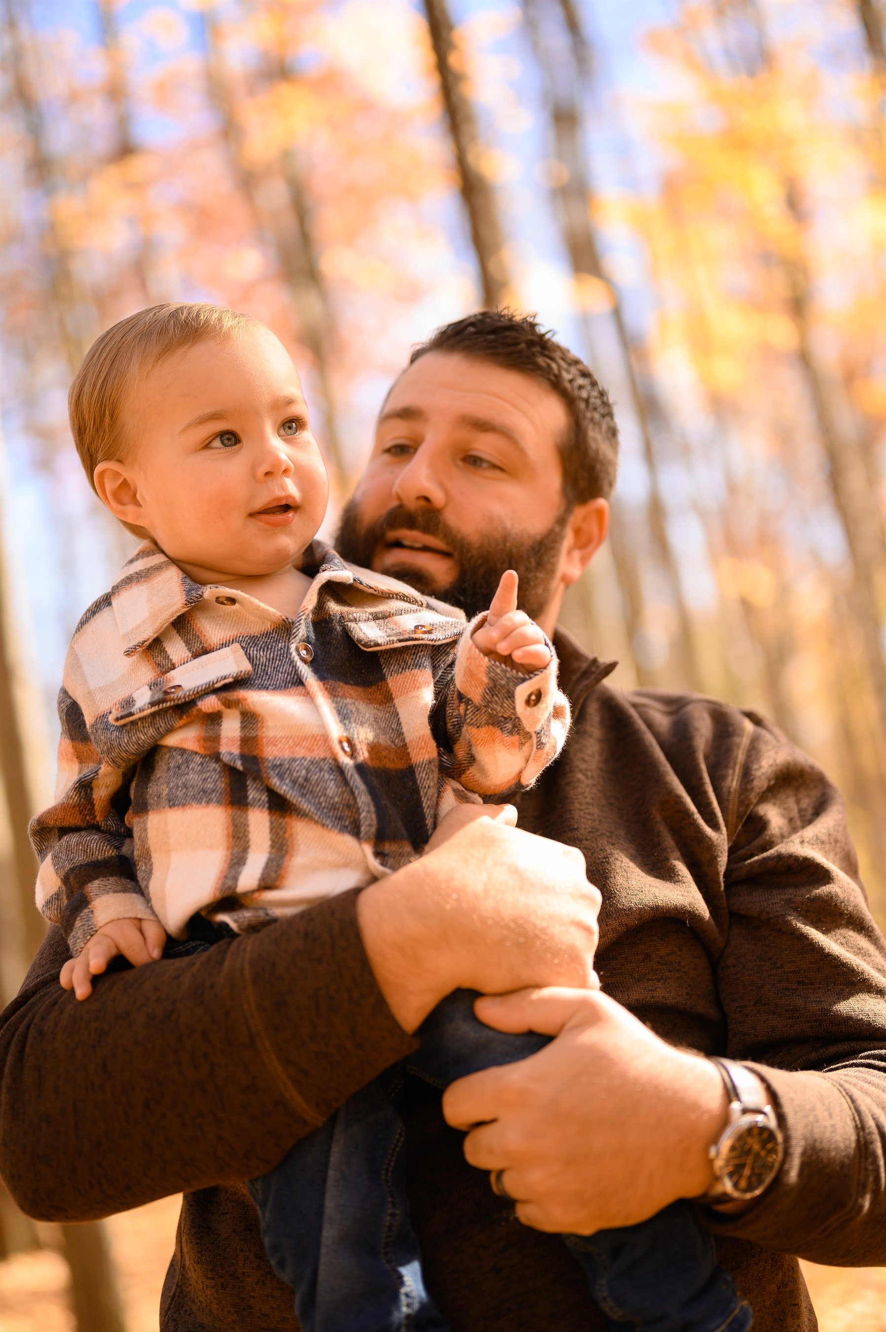 Man holding a baby in a plaid shirt, standing in a forest with autumn leaves.