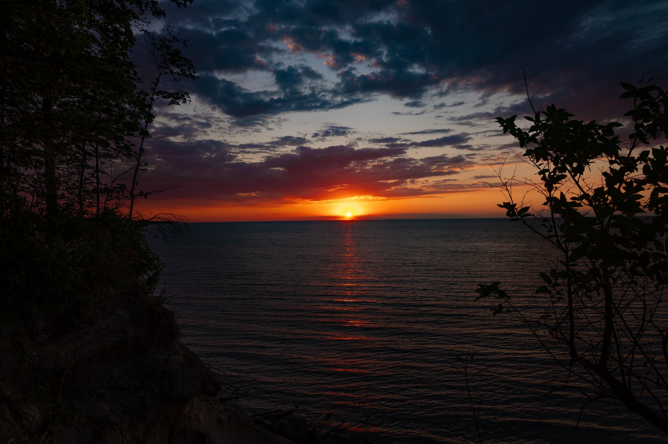 Sunset over a calm ocean with silhouetted trees and colorful clouds.