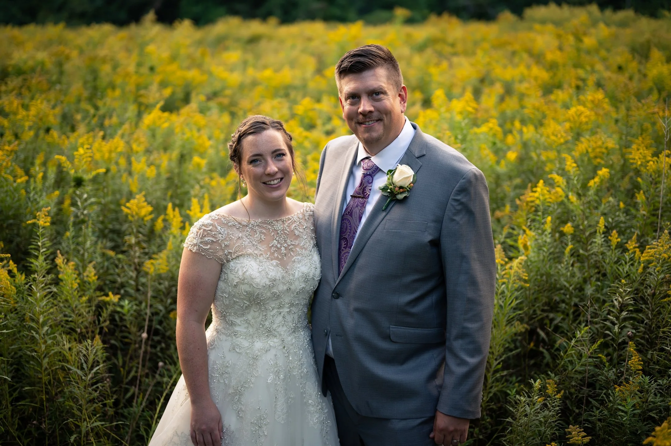 Bride and groom in field of yellow flowers, wearing wedding attire.