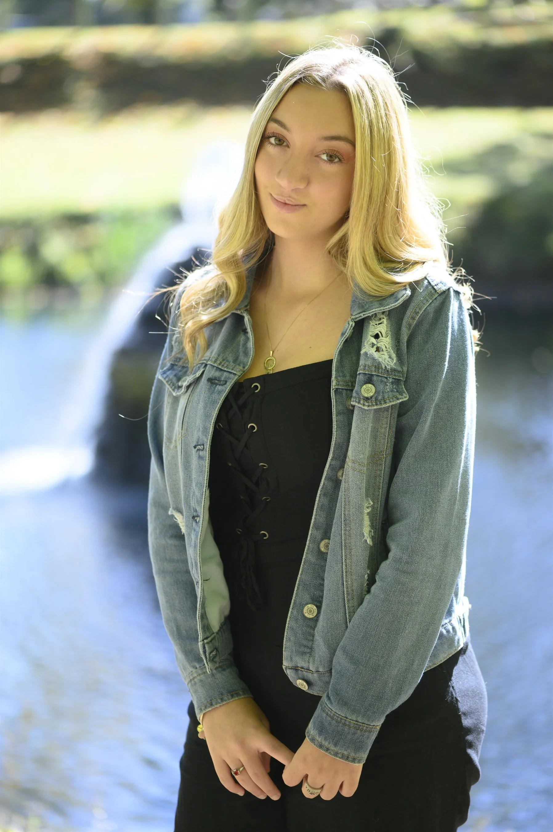 Person with long blonde hair wearing a denim jacket and black dress standing outdoors near water.