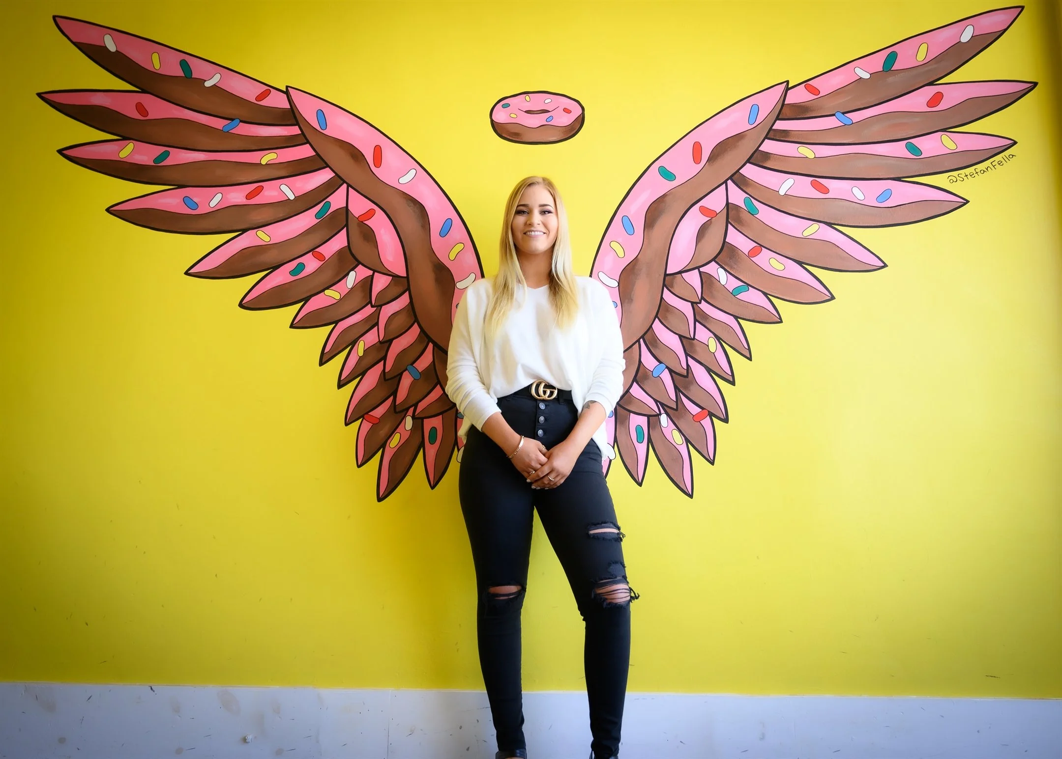 A woman standing in front of a bright yellow wall, where there is a mural of pink and brown doughnut-shaped wings with colorful sprinkles, giving the appearance that she has wings. The mural also features a small doughnut halo above her head.