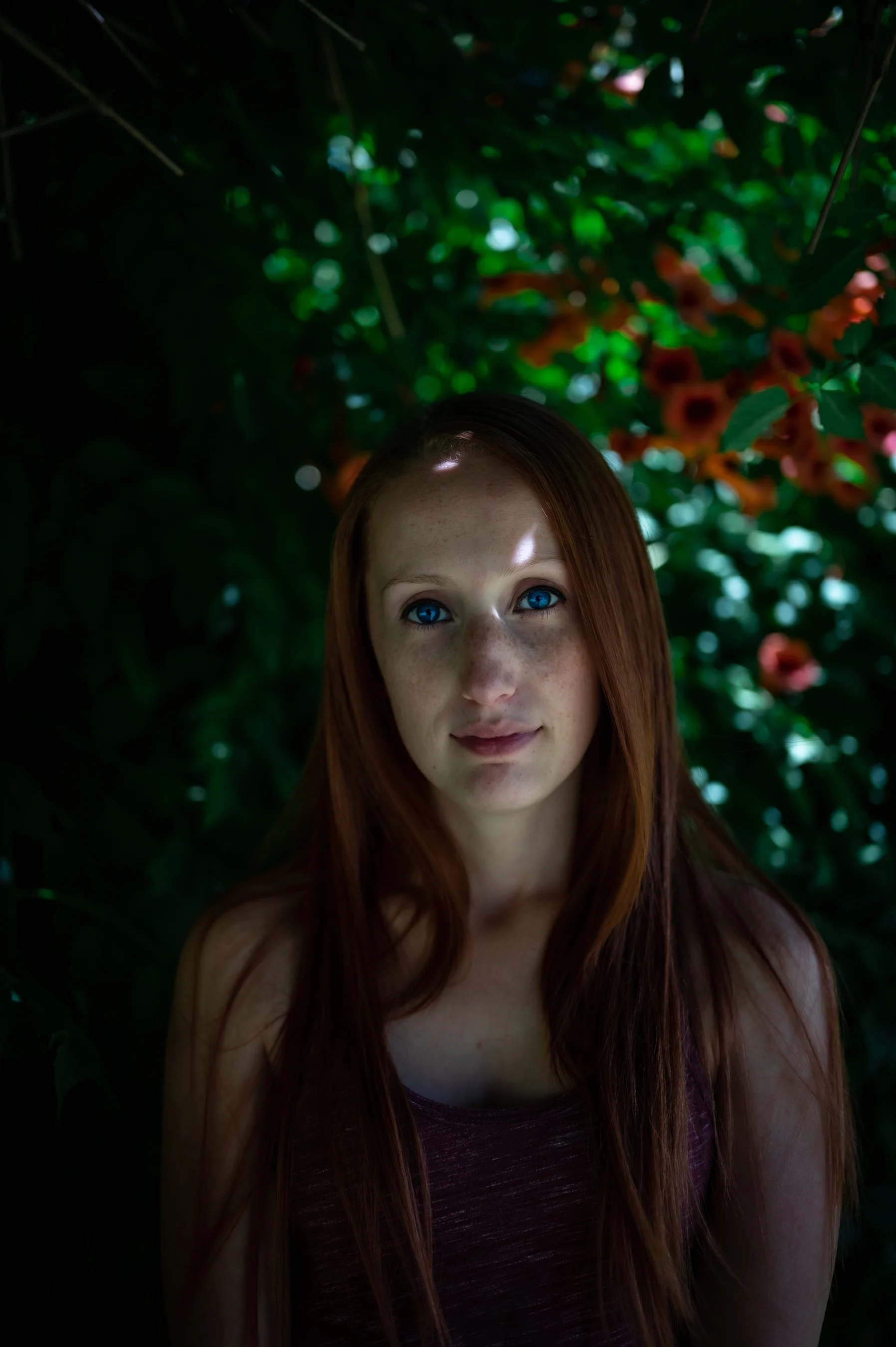 Portrait of a woman with long red hair and blue eyes, standing in front of lush green foliage with orange flowers.