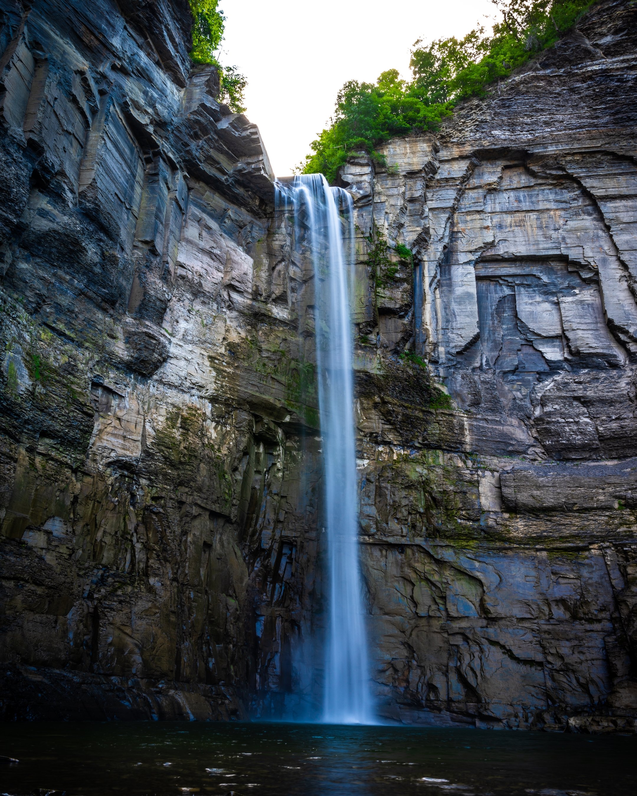 Tall waterfall cascading down rocky cliffs into a pool, surrounded by lush green vegetation.
