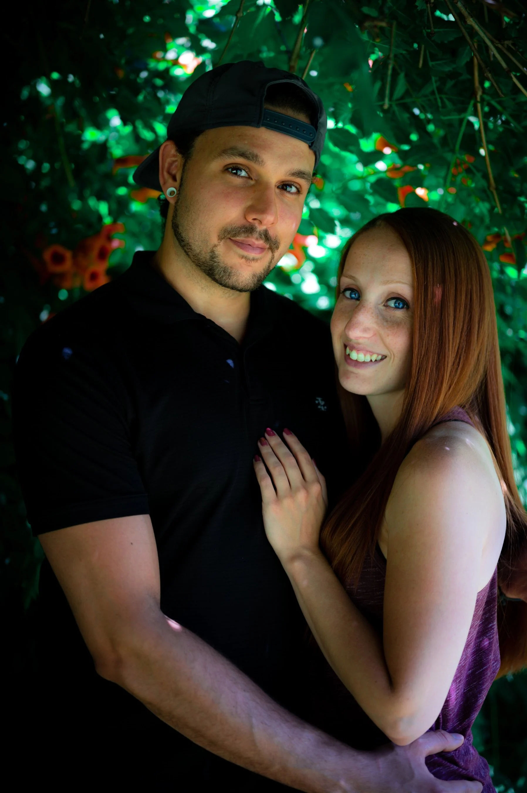 A couple smiling and embracing in a garden with green foliage and flowers in the background.